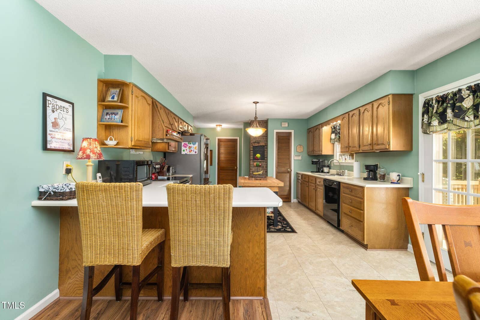A spacious kitchen with teal walls, wooden cabinets, a white countertop island with two wicker bar stools, and modern appliances. Sunlight enters through glass doors on the right, and a dining table is partially visible.