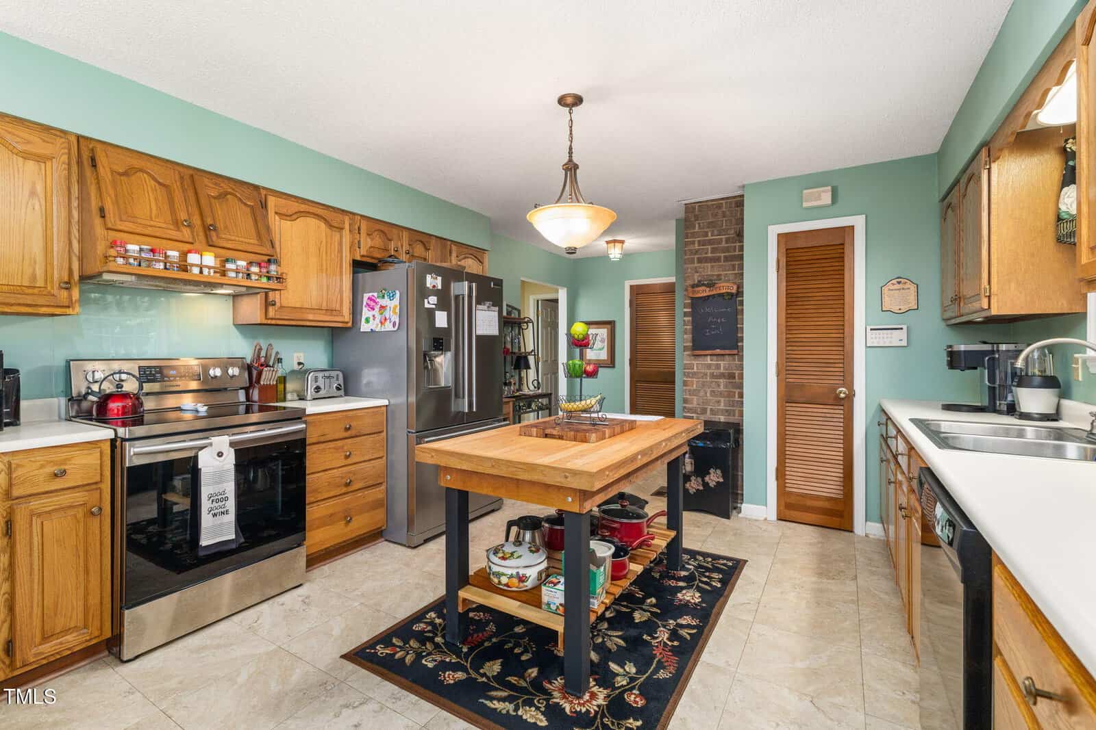 A kitchen with wooden cabinets, stainless steel appliances, a central island with a fruit bowl, green walls, and tile flooring. A rug sits under the island, and various kitchen items are arranged on counters and shelves.
