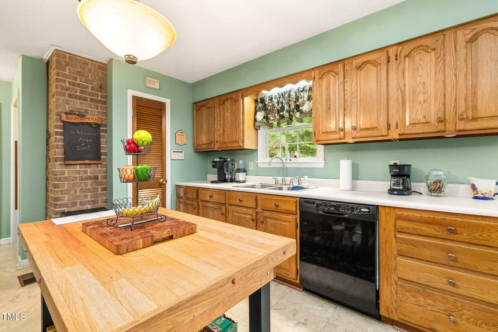 A kitchen with wooden cabinets, white countertops, a black dishwasher, and a window above the sink. An island with a wooden top holds a fruit basket. There is a brick accent wall and a chalkboard in the background.