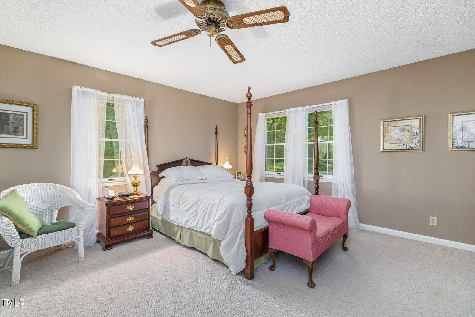 A cozy bedroom with a four-poster bed, pink bench, wicker chair, wooden nightstand, and two windows with white curtains. Ceiling fan overhead and framed art on tan walls.
