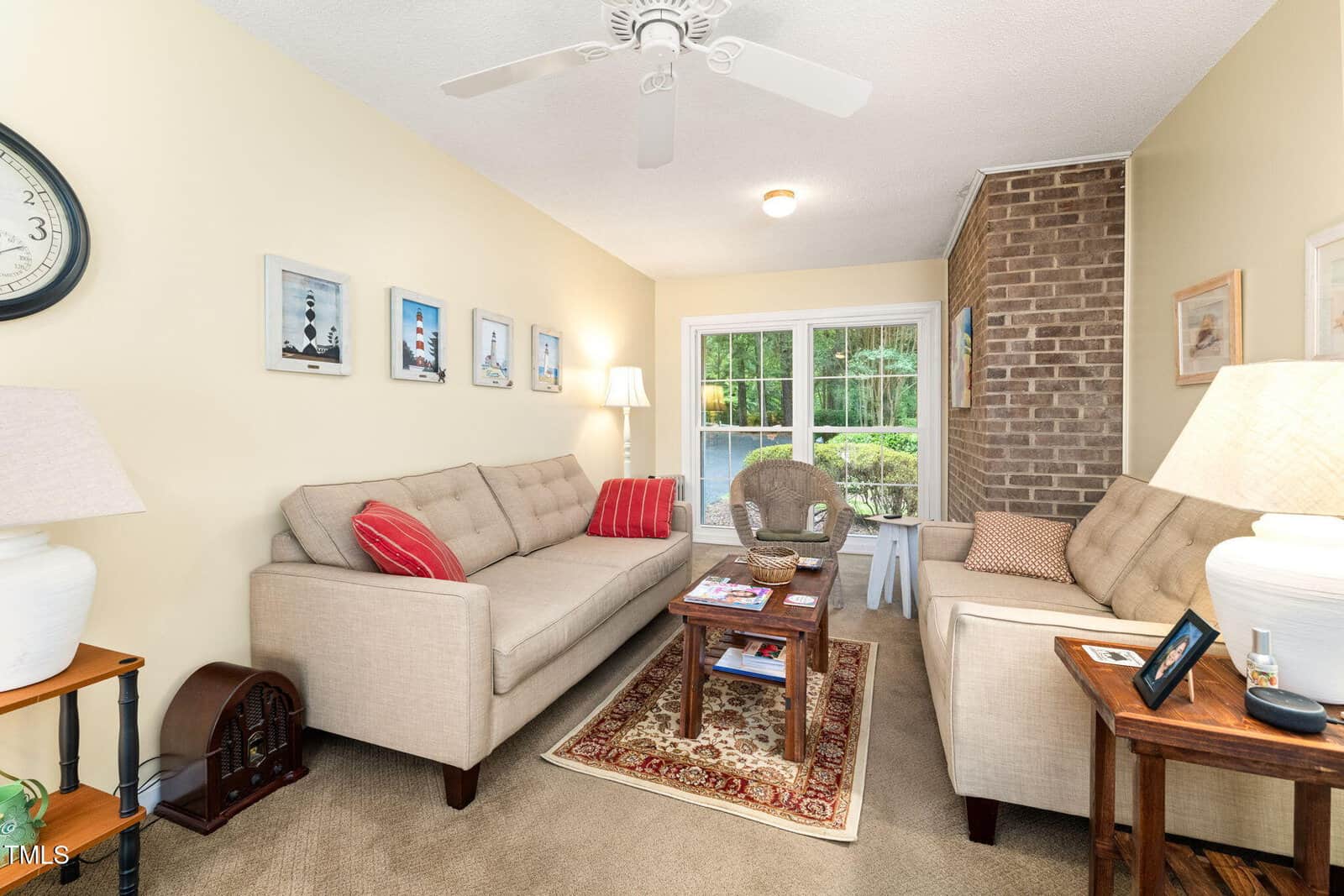 Cozy living room with beige sofas, colorful pillows, a small rug, lamps, side tables, artwork on the wall, large window showing greenery, and a brick accent wall in the corner.