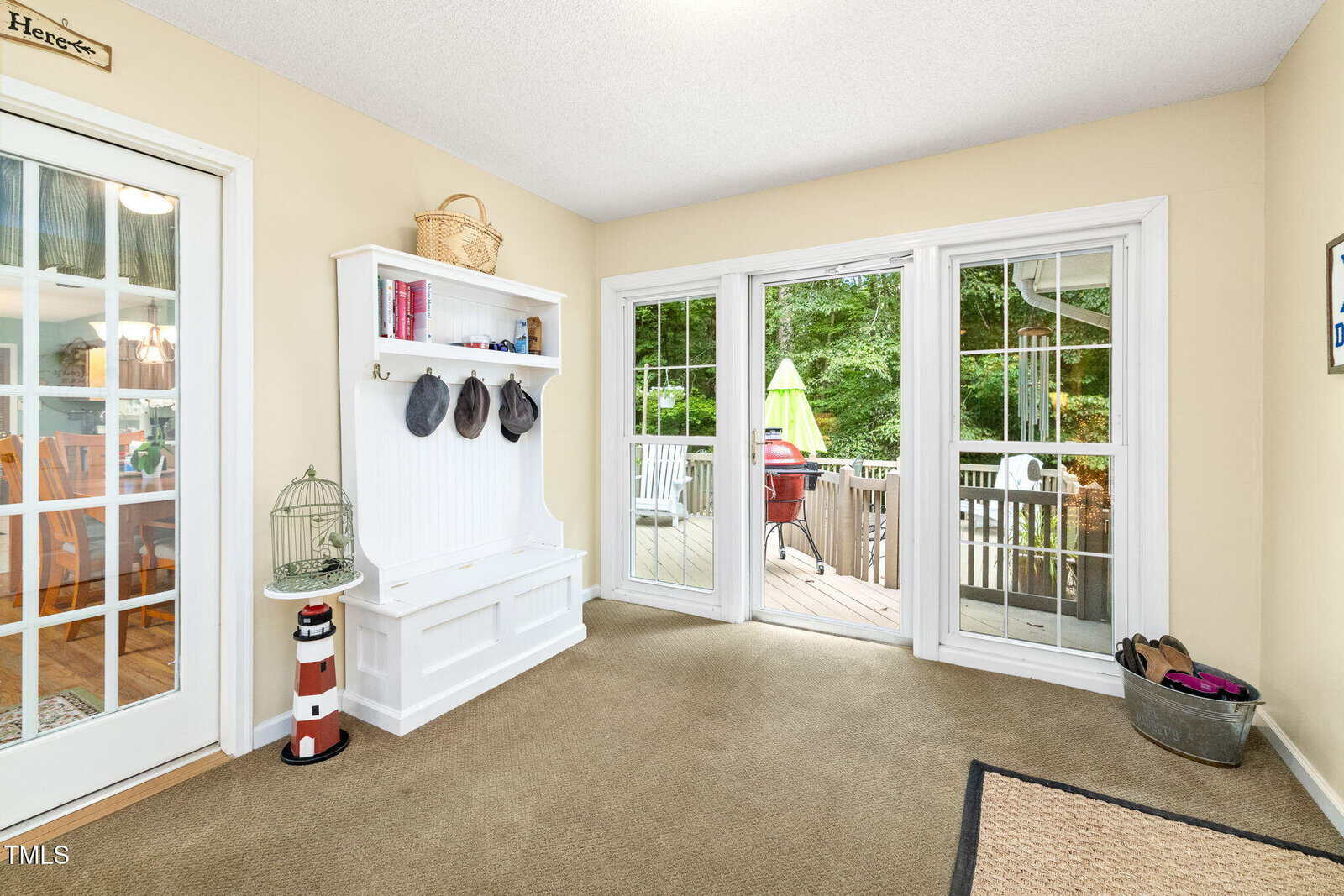 Bright sunroom with beige walls, a white bench with hooks, and double glass doors leading to a deck with outdoor furniture. A birdcage, hats, books, and shoes add casual decorative touches.