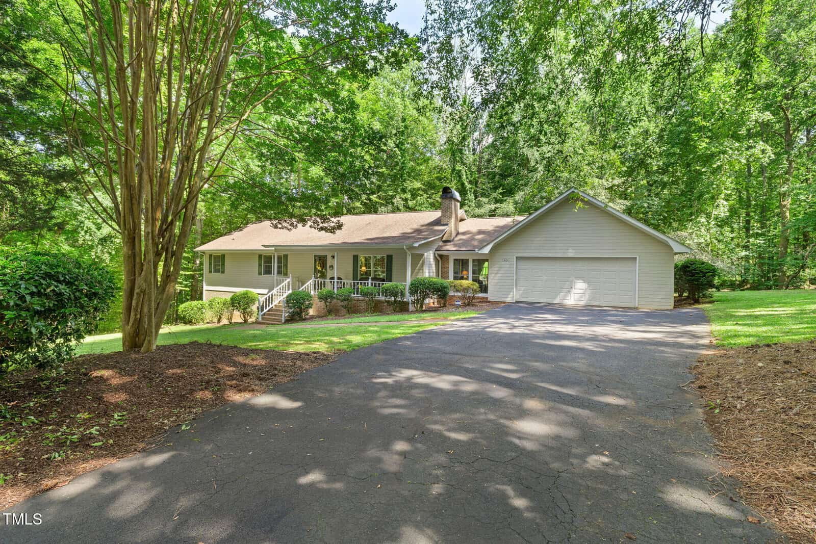 Charming single-story Oxford home at 5204 Tabbs Creek Lane featuring a front porch, attached three-car garage, and dormer windows, nestled among trees and greenery with a paved driveway leading up to the residence.
