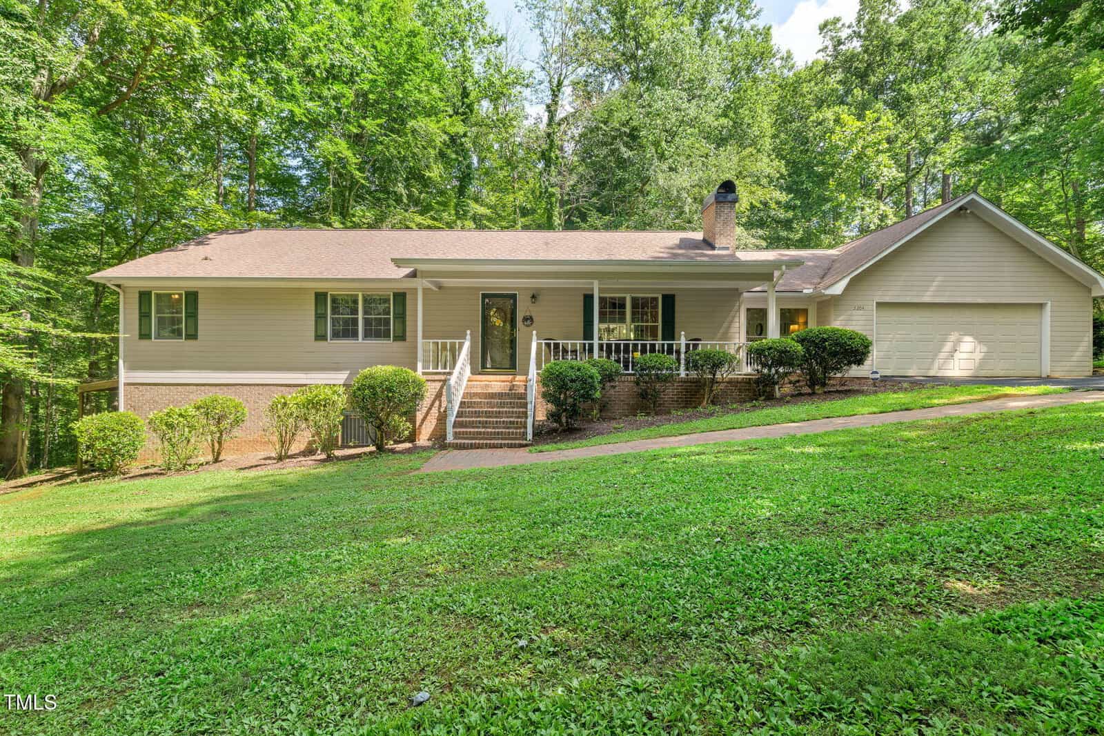 Charming single-story beige house at 5204 Tabbs Creek Lane, Oxford, featuring a covered front porch, brick steps, multiple windows, and an attached two-car garage on a grassy lawn with shrubs and tree-lined surroundings.