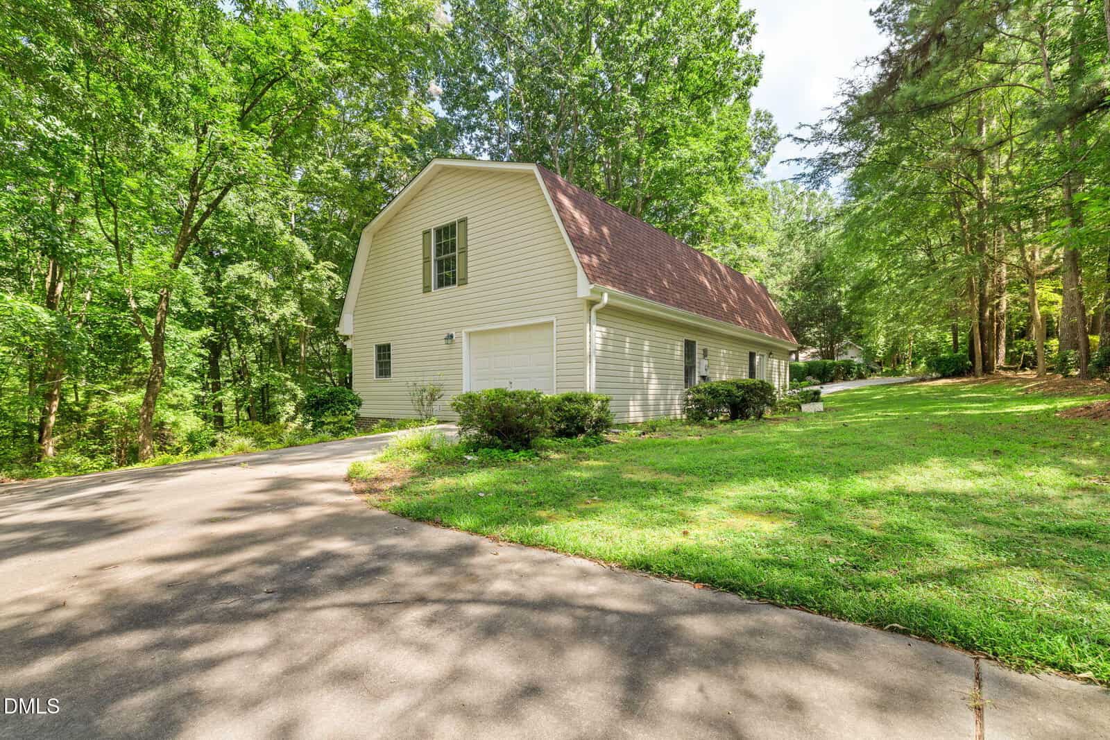 A two-story beige house with a gambrel roof sits next to a curved driveway, surrounded by green grass, bushes, and tall trees in a wooded area on a sunny day.