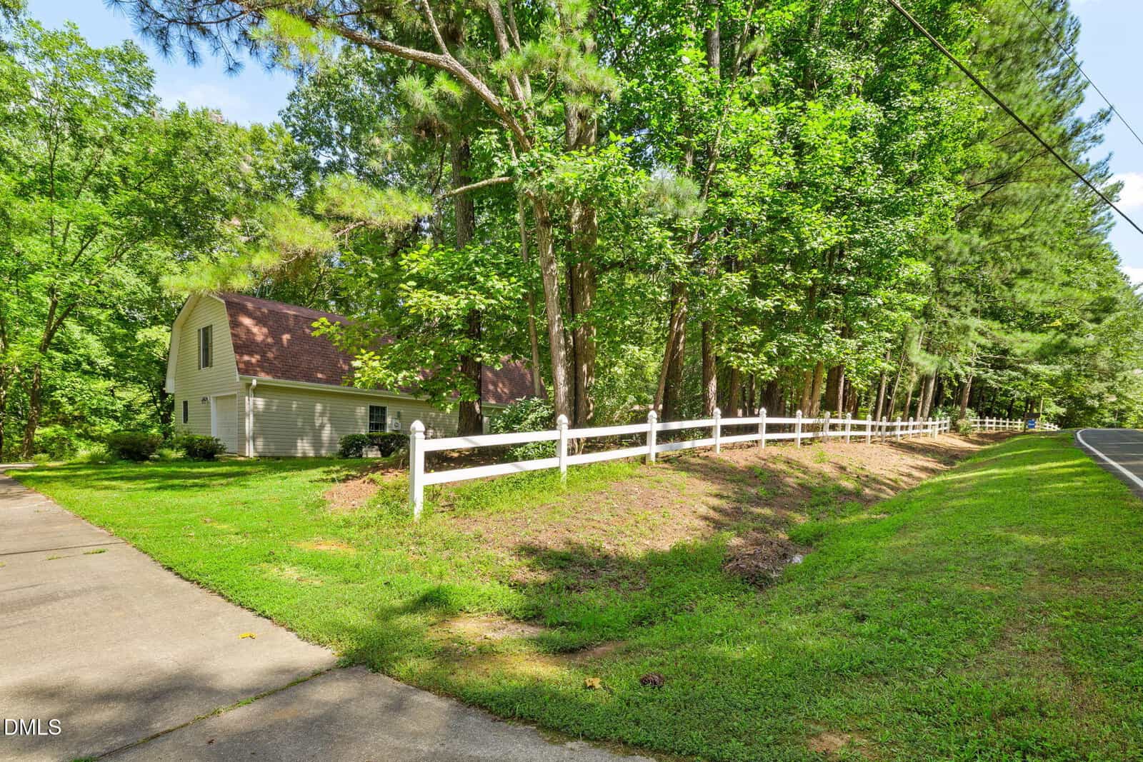 A white house with a brown roof sits behind a white wooden fence, surrounded by tall green trees and grass. A paved driveway and a two-lane road run along the right side of the property.