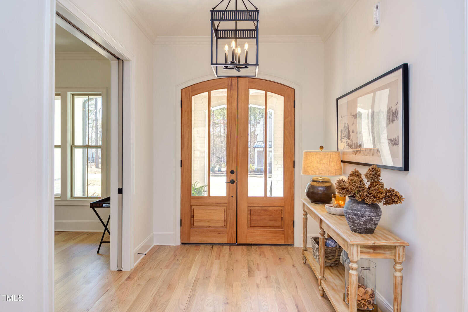 A bright entryway with wooden double doors, light hardwood floors, a lantern-style ceiling light, a console table with a lamp, and decorative vases. A large framed photo hangs on the wall above the table.