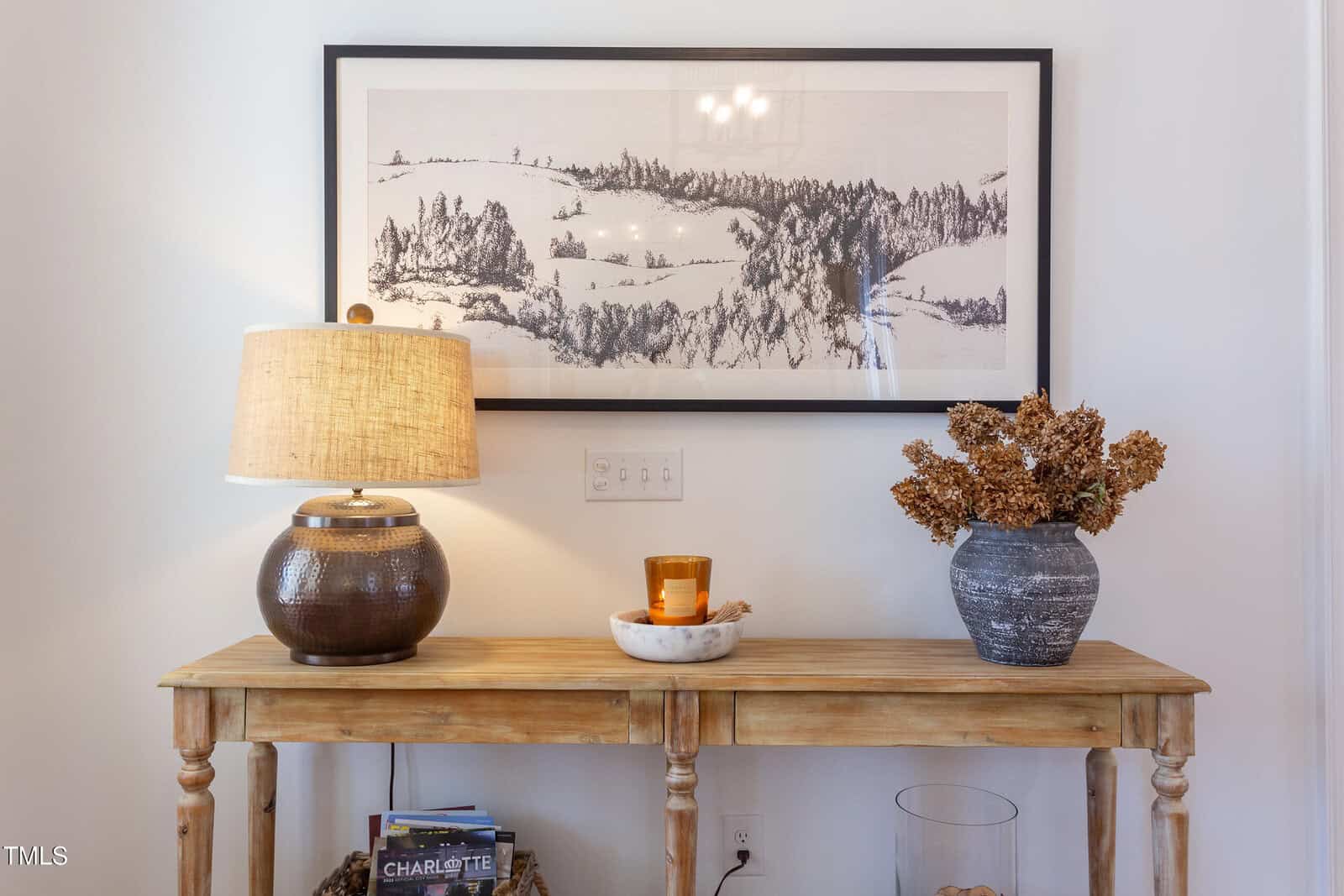A wooden console table against a white wall holds a brown lamp, a candle on a marble tray, and a vase with dried flowers. A framed landscape art print hangs above the table. Magazines are on the lower shelf.