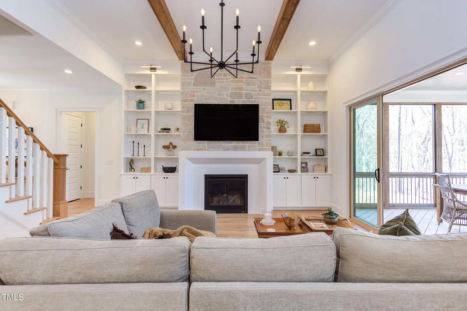 A living room with a beige sectional sofa facing a fireplace with a mounted TV, built-in shelves on both sides, wood ceiling beams, a large black chandelier, and sliding glass doors leading to a screened porch.
