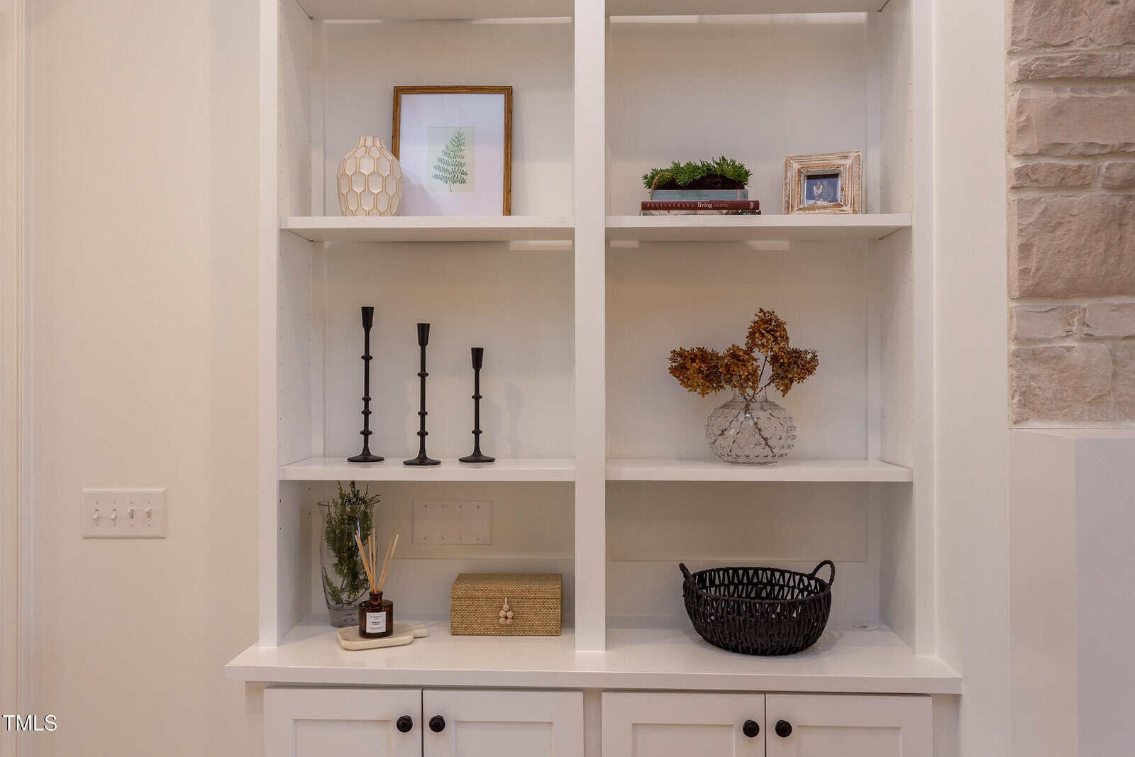 A white built-in shelving unit displays framed art, decor items, candlesticks, a vase with dried flowers, a small plant, a wicker basket, and various decorative objects on open shelves above closed cabinets.