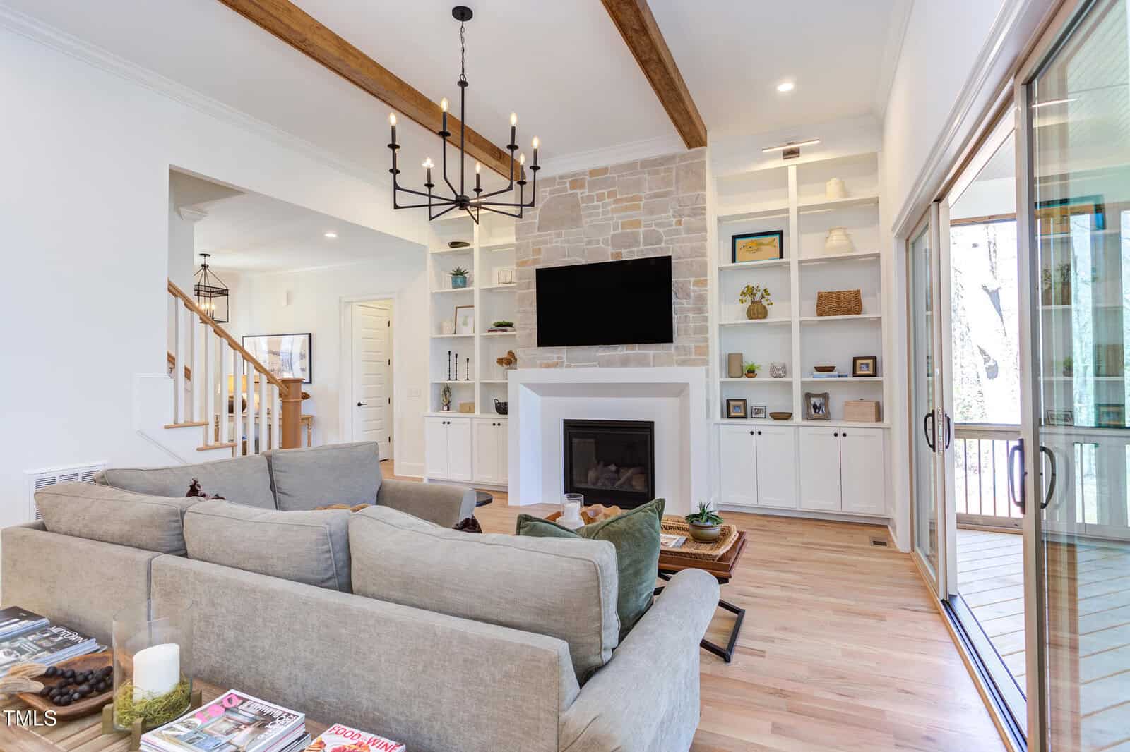 A modern living room with light wood floors, beige sectional sofa, a stone fireplace with a TV above it, built-in shelves, large windows, and exposed wooden ceiling beams. A staircase is visible to the left.