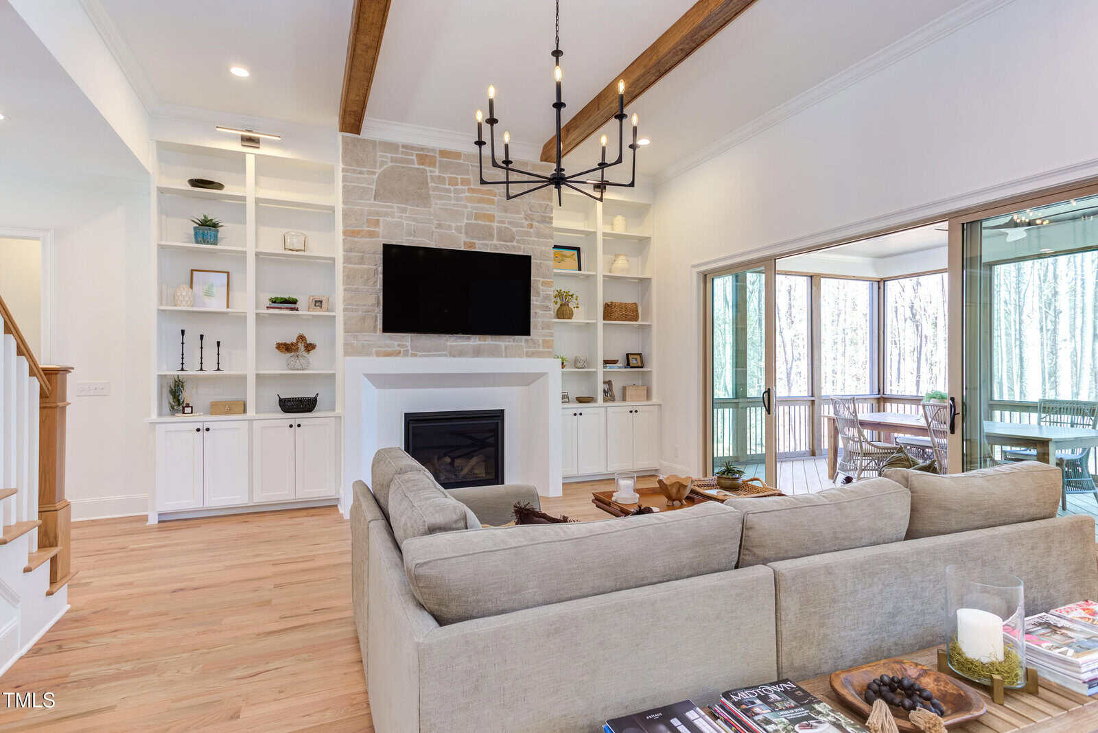 Bright living room with a beige sectional sofa, stone fireplace, mounted TV, built-in shelves, wooden beams, chandelier, and sliding glass doors opening to a screened porch with forest view.