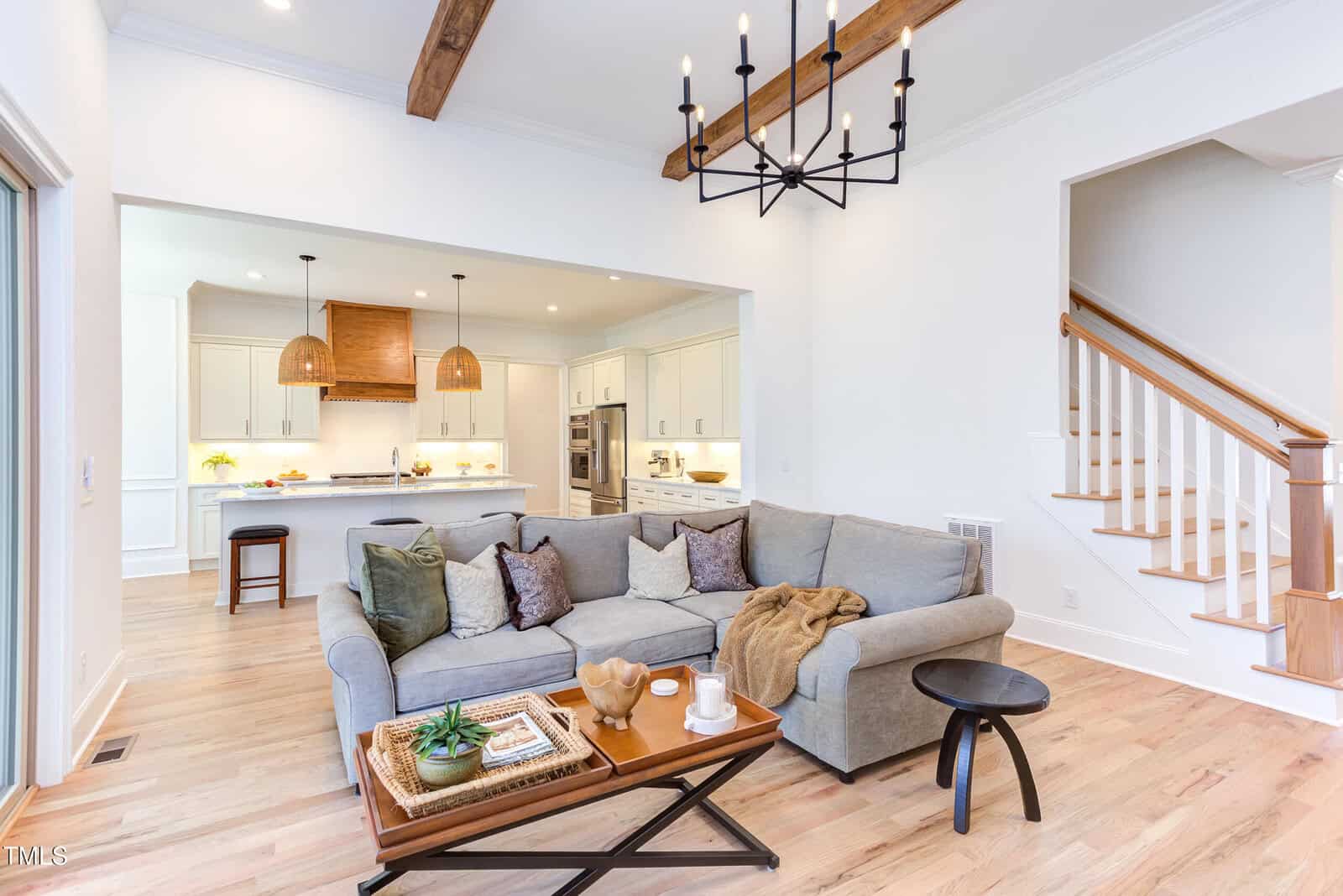 Bright living room with a gray sectional sofa, wooden coffee table, and throw blankets, open to a modern white kitchen with pendant lights. Wooden beams and staircase are visible.