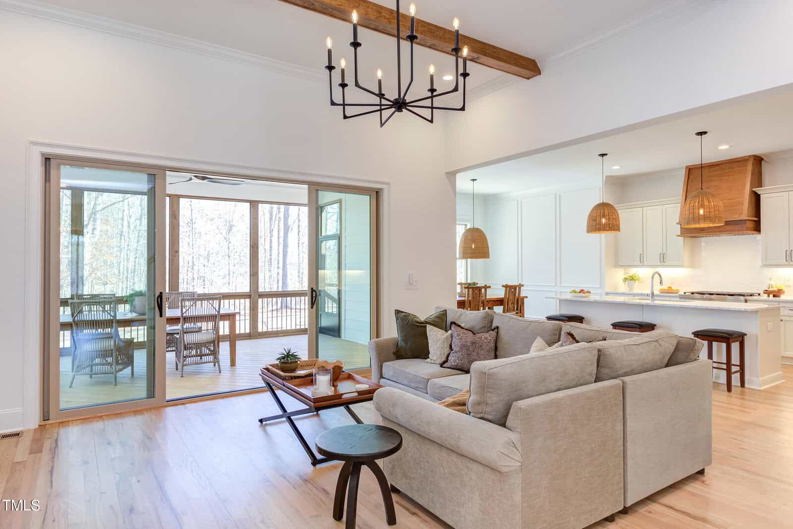 Bright living room with a gray sectional sofa, wooden coffee table, and modern chandelier, open to a white kitchen and an adjoining sunroom with large glass sliding doors showing a wooded view outside.