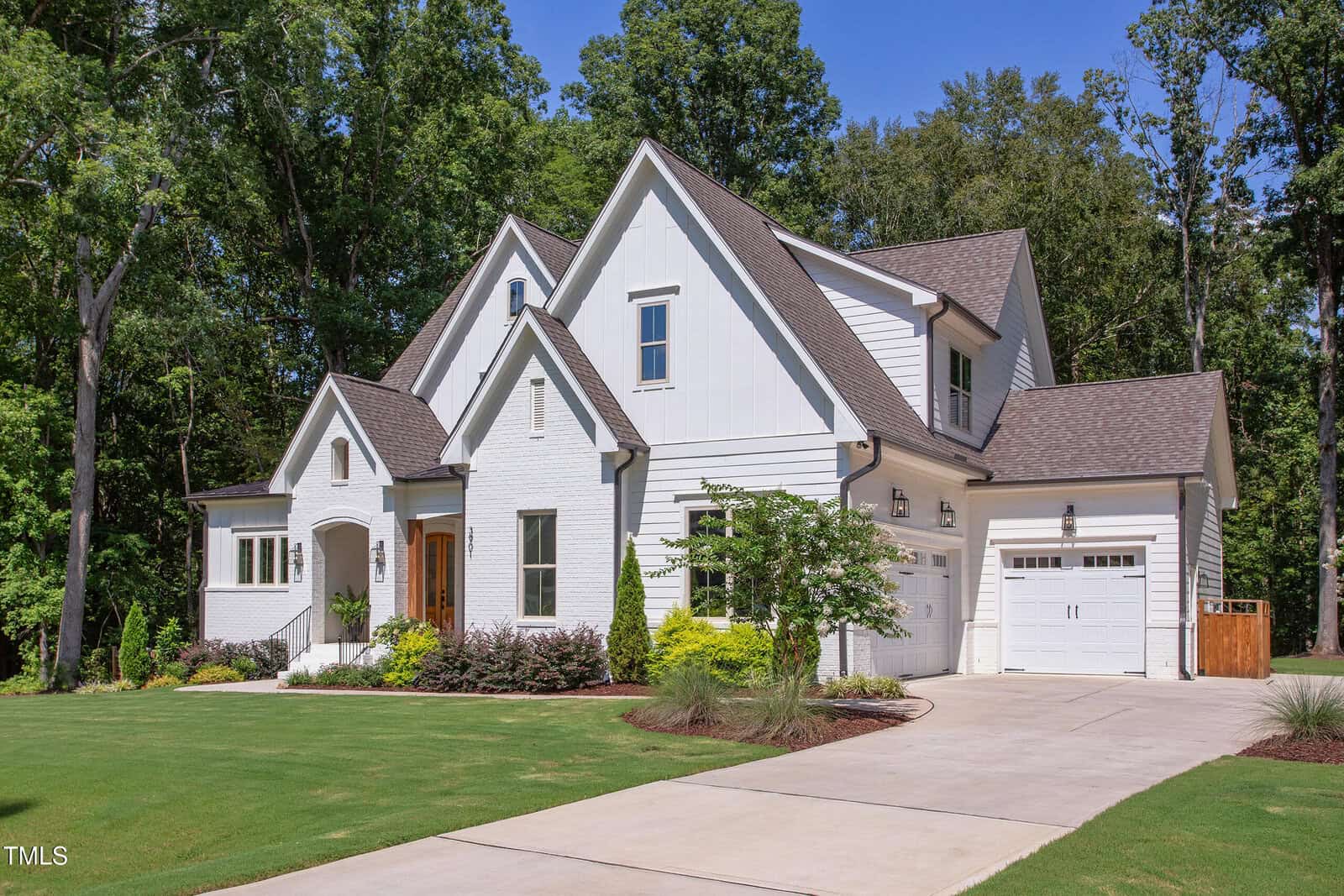 A two-story white modern farmhouse with brick and siding exterior, gabled roofs, a double garage, manicured lawn, and landscaped plants, set against tall trees and a clear blue sky.