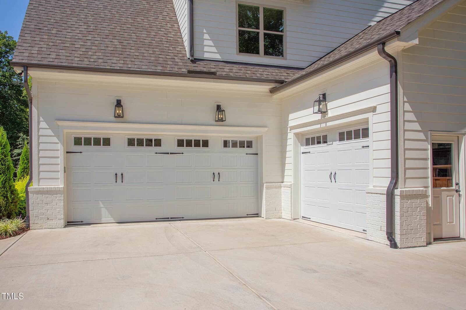 Two white garage doors and a side entry door on a modern house exterior with white siding, brick trim, outdoor lights, and a clean concrete driveway.