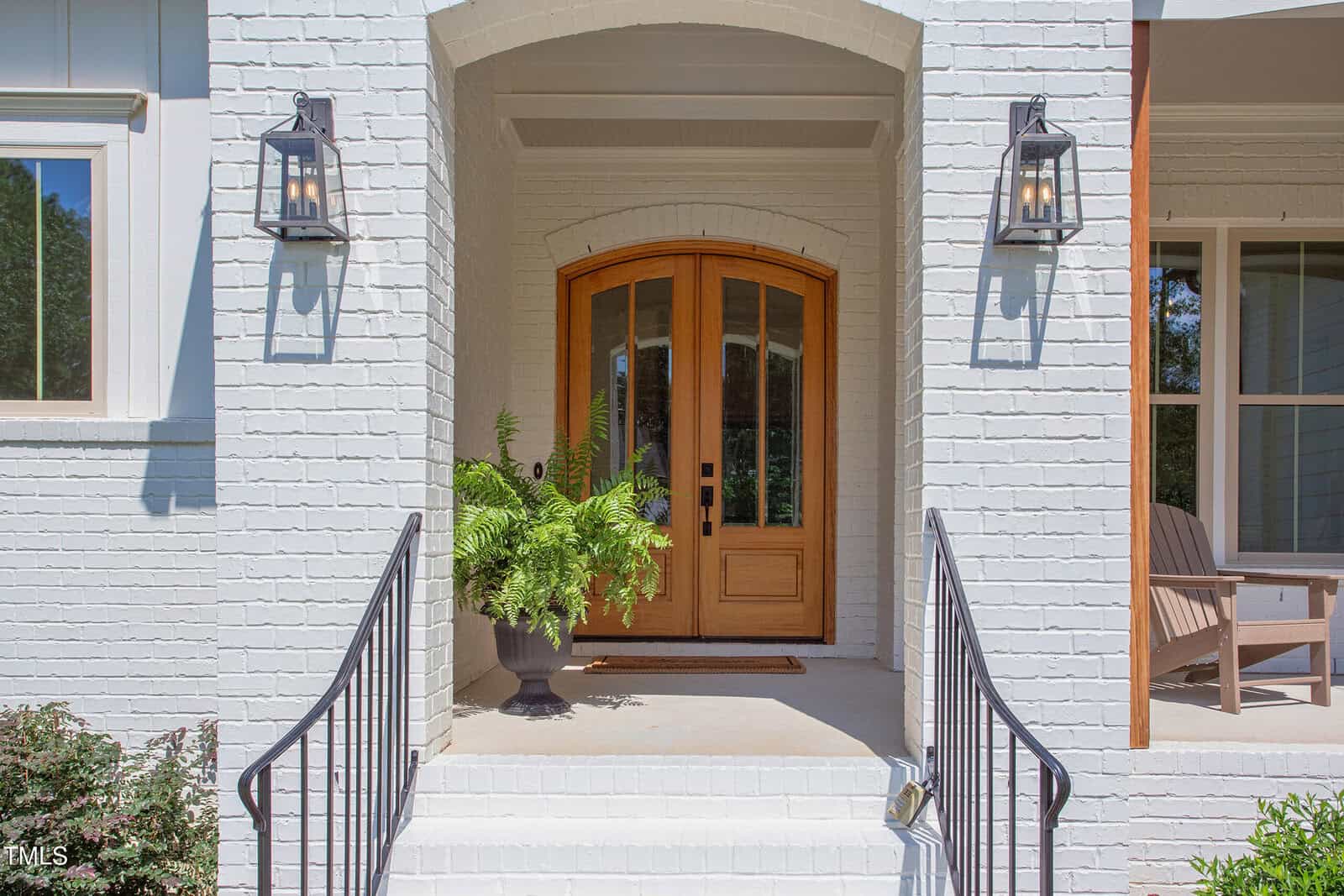 A wooden double front door with glass panels is centered on a white brick porch. Two wall-mounted lanterns flank the door, and a potted fern sits to the left. Black metal railings line the steps leading up to the entry.