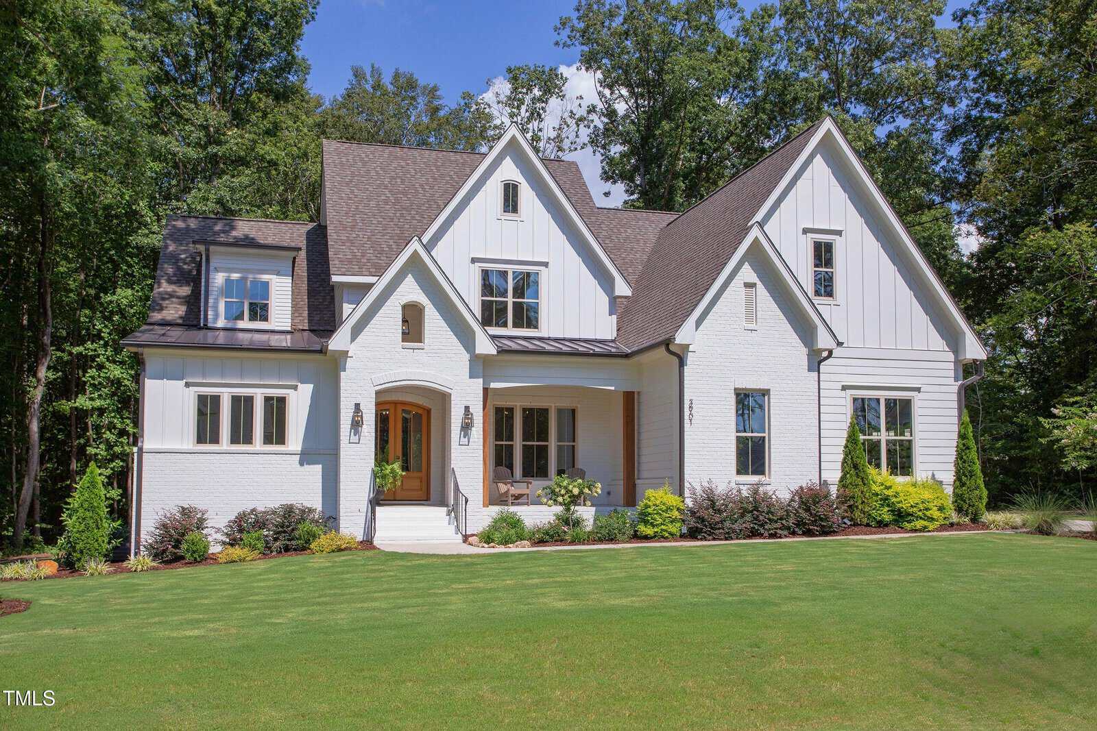 A two-story white house with a brown shingled roof, wood front door, multiple gables, and large windows, surrounded by green grass, shrubs, and trees under a clear blue sky.