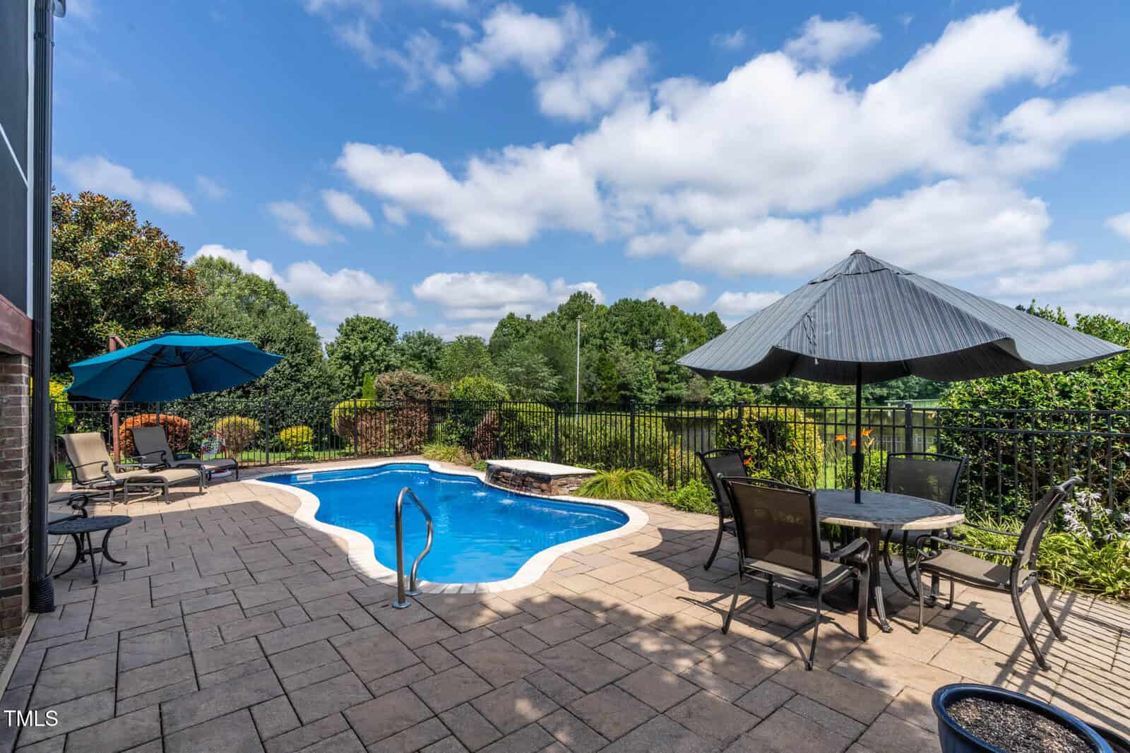 Outdoor patio with a small in-ground pool, surrounded by lounge chairs, tables with umbrellas, and a landscaped garden. A metal fence encloses the area, and trees fill the background under a partly cloudy sky.