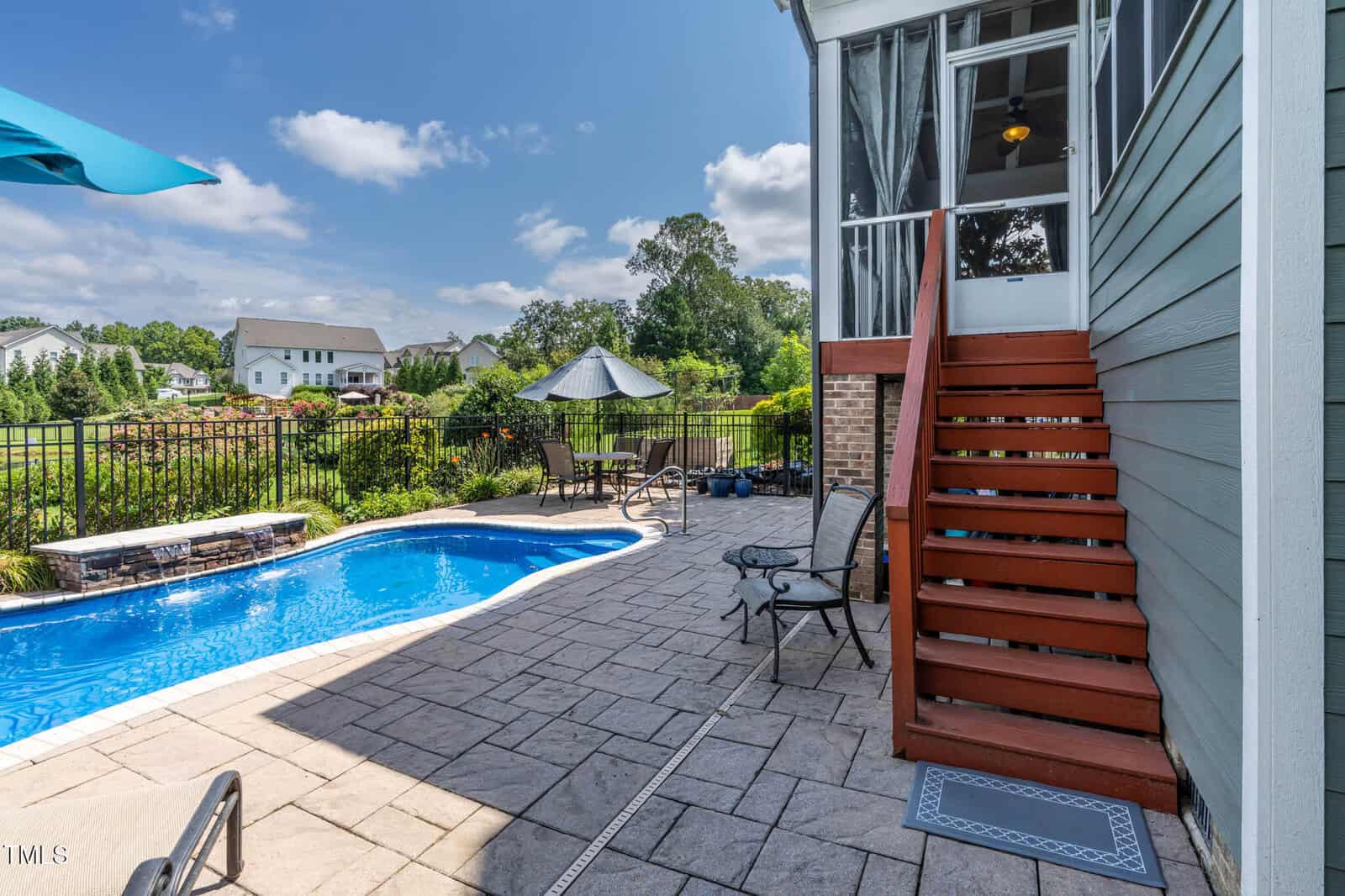 Outdoor scene with a blue swimming pool, stone patio, metal fence, table with umbrella, lounge chairs, and red wooden stairs leading to a screened porch attached to a house. Other houses and greenery are visible in the background.
