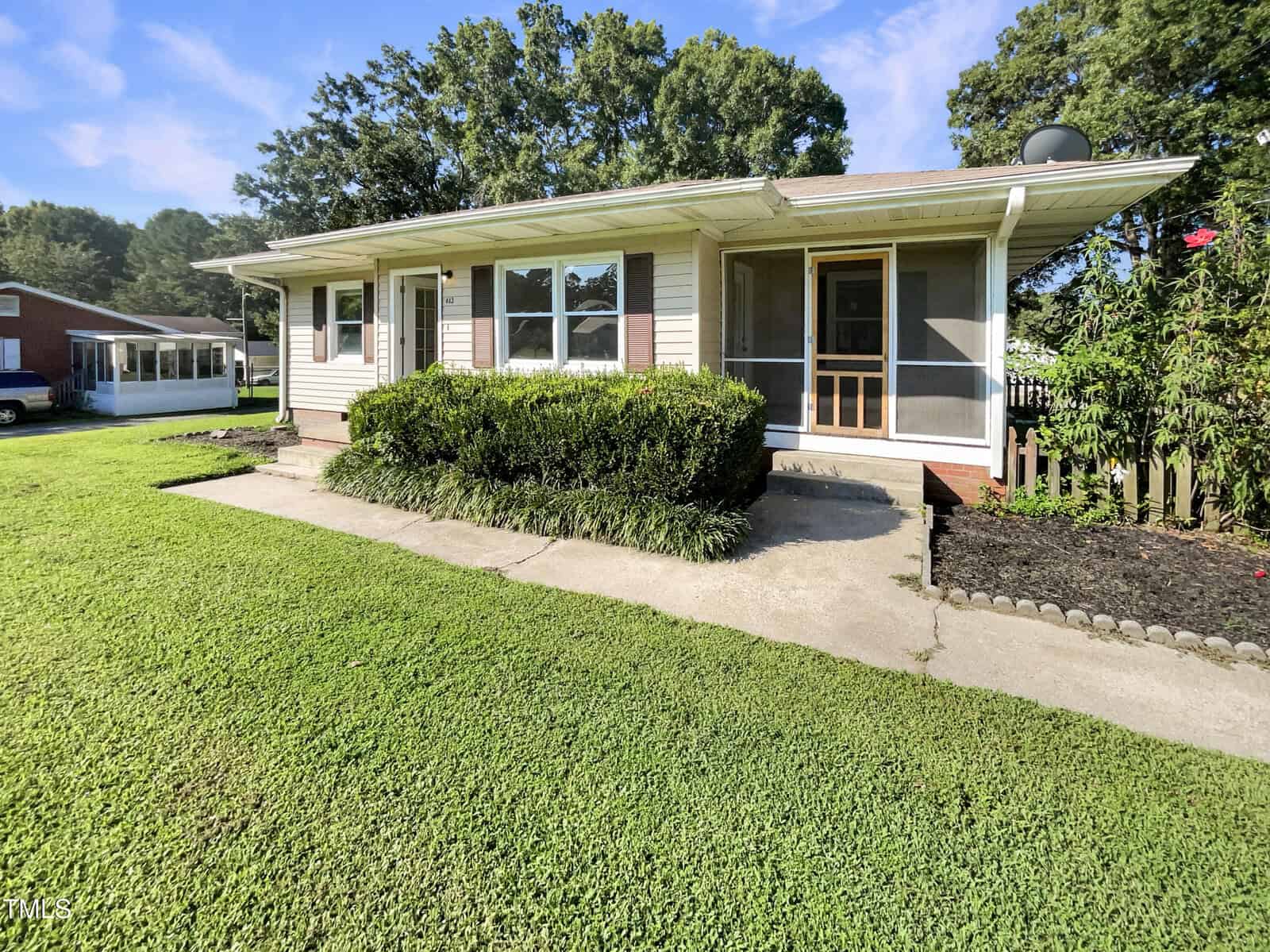 Charming single-story home at 613 15th Street, Butner, featuring beige siding with white trim, a screened front door, multiple windows, concrete walkway, trimmed hedges, and a well-maintained lawn under a blue sky with scattered clouds.