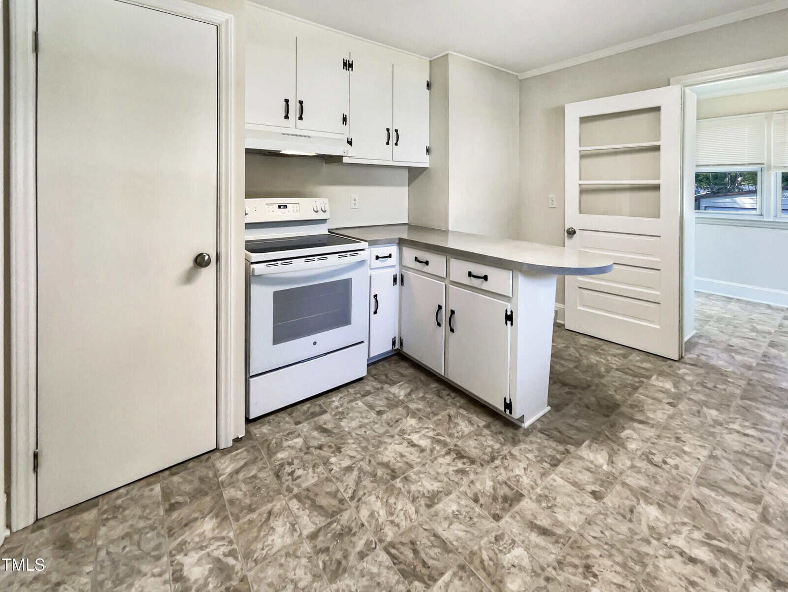 A kitchen with white cabinets, a white electric stove, and a small peninsula counter. The floor is patterned vinyl, and there are doors leading to other rooms and a set of built-in shelves.