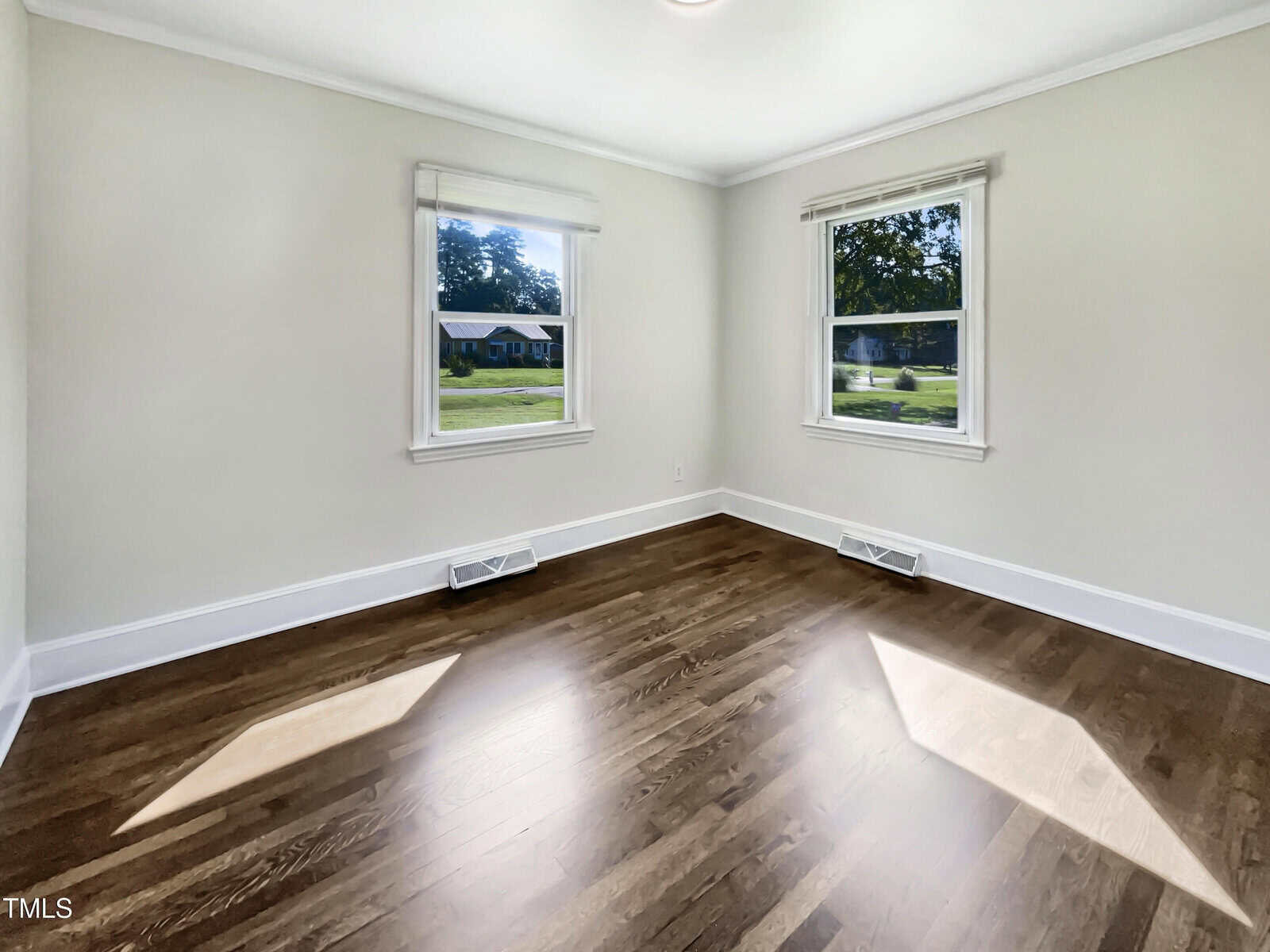 Empty room with light-colored walls, two windows with white trim, hardwood floors, white baseboards, and two floor vents beneath the windows. Sunlight enters through the windows, casting bright patches on the floor.