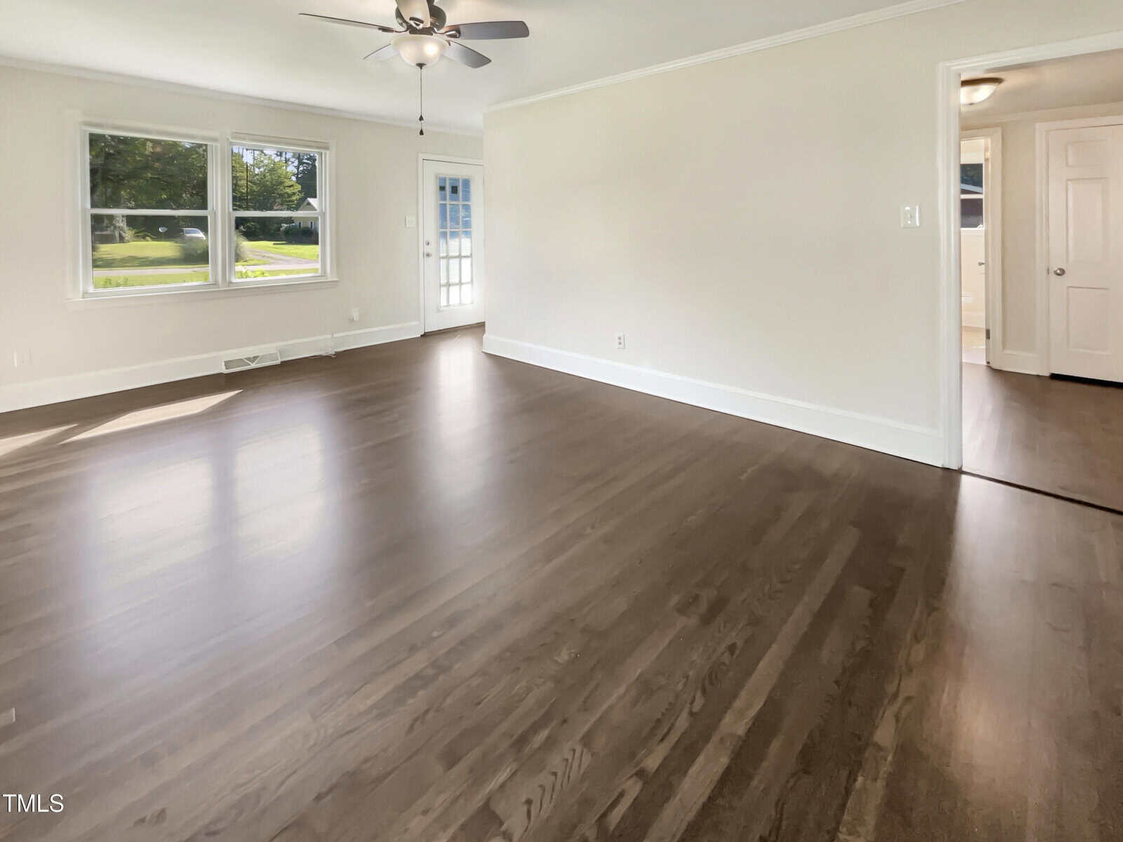 An empty room with polished hardwood floors, beige walls, a ceiling fan with lights, three windows letting in natural light, and a door leading to another room.