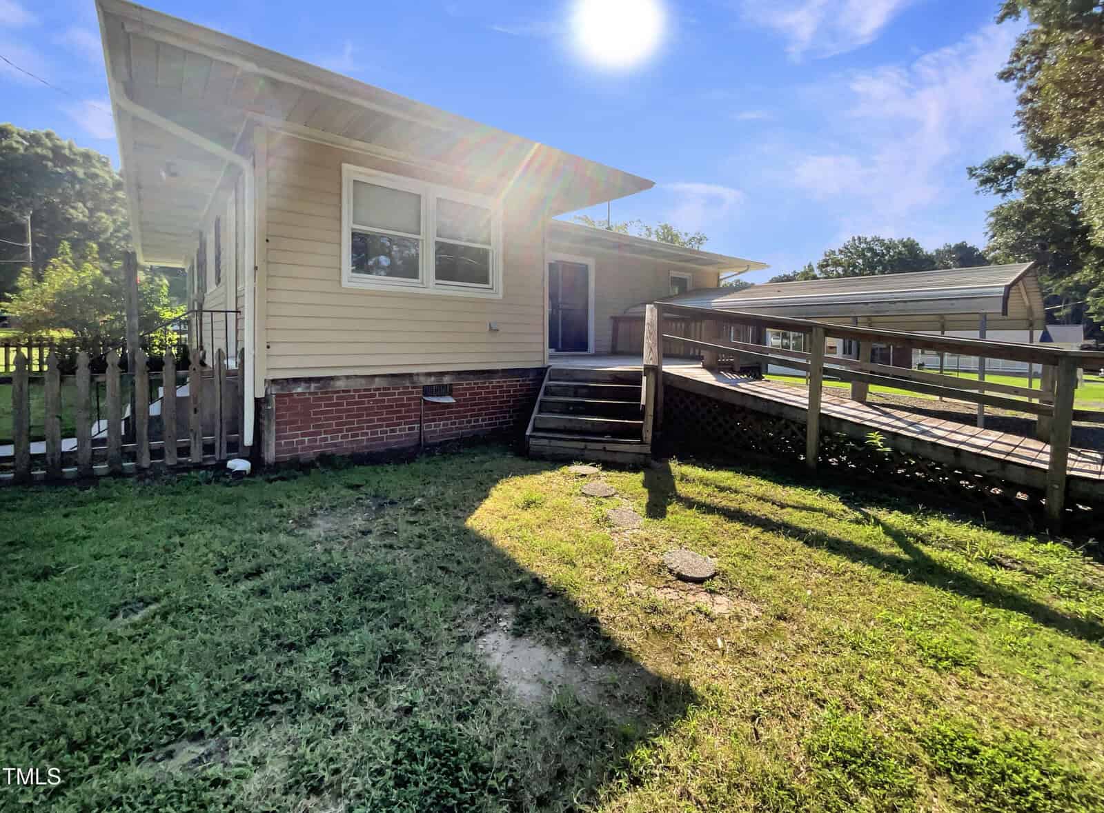 Single-story beige house with a brick foundation, wooden ramp, steps, and fenced area. The sun is bright in a clear blue sky, casting shadows on the grassy yard. Other homes and trees are in the background.