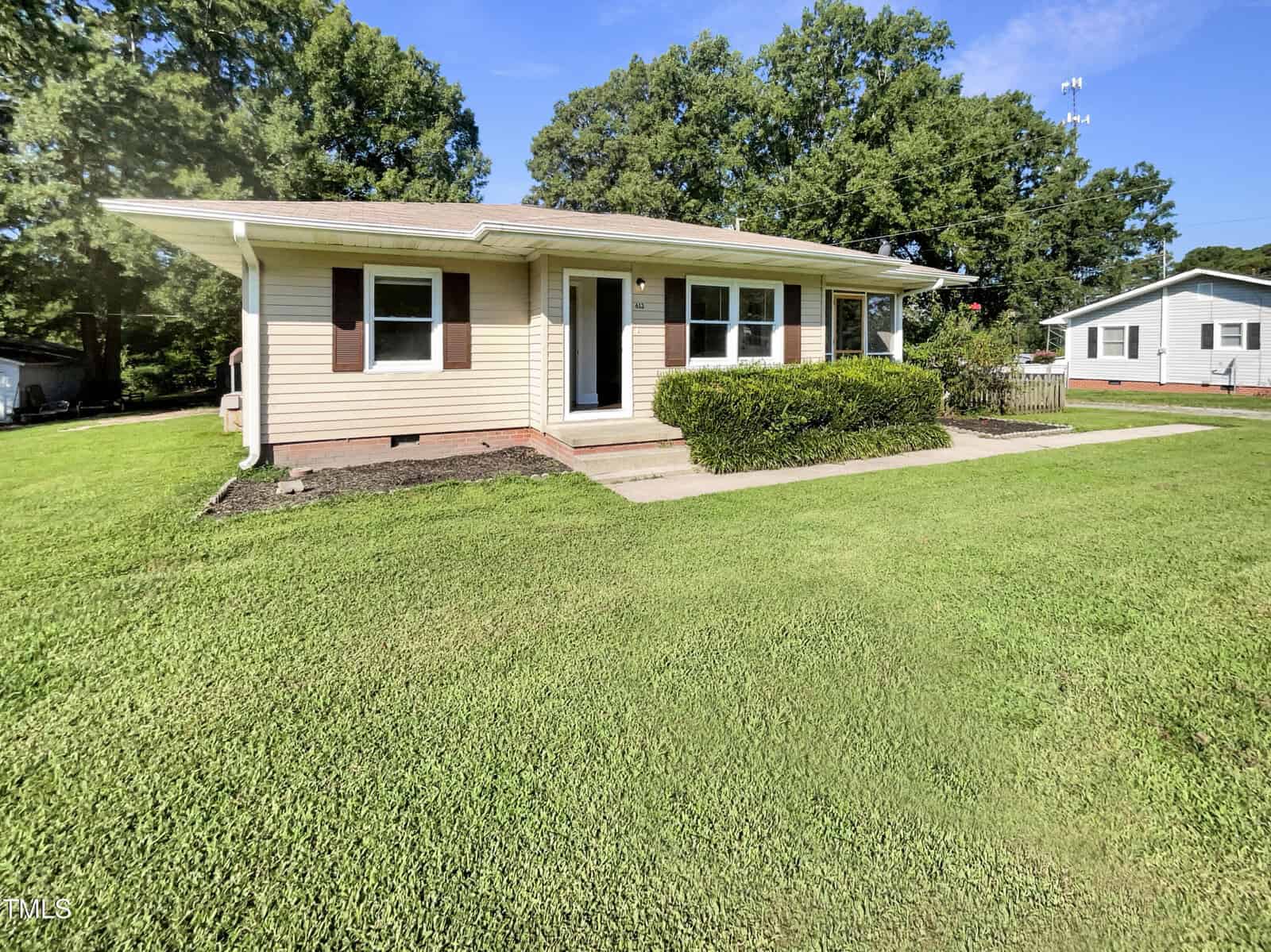 Single-story house with tan siding, brown shutters, and a covered front porch. The house is surrounded by green grass and shrubs, with a concrete walkway leading to the front door. Trees and another house are in the background.