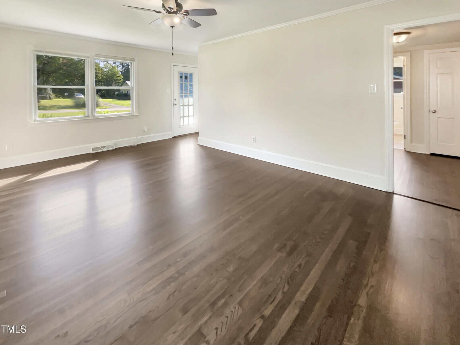 Empty room with light-colored walls, large windows, hardwood floors, a ceiling fan with light, and a door leading outside. An open doorway reveals a hallway and additional room. Sunlight enters through the windows.