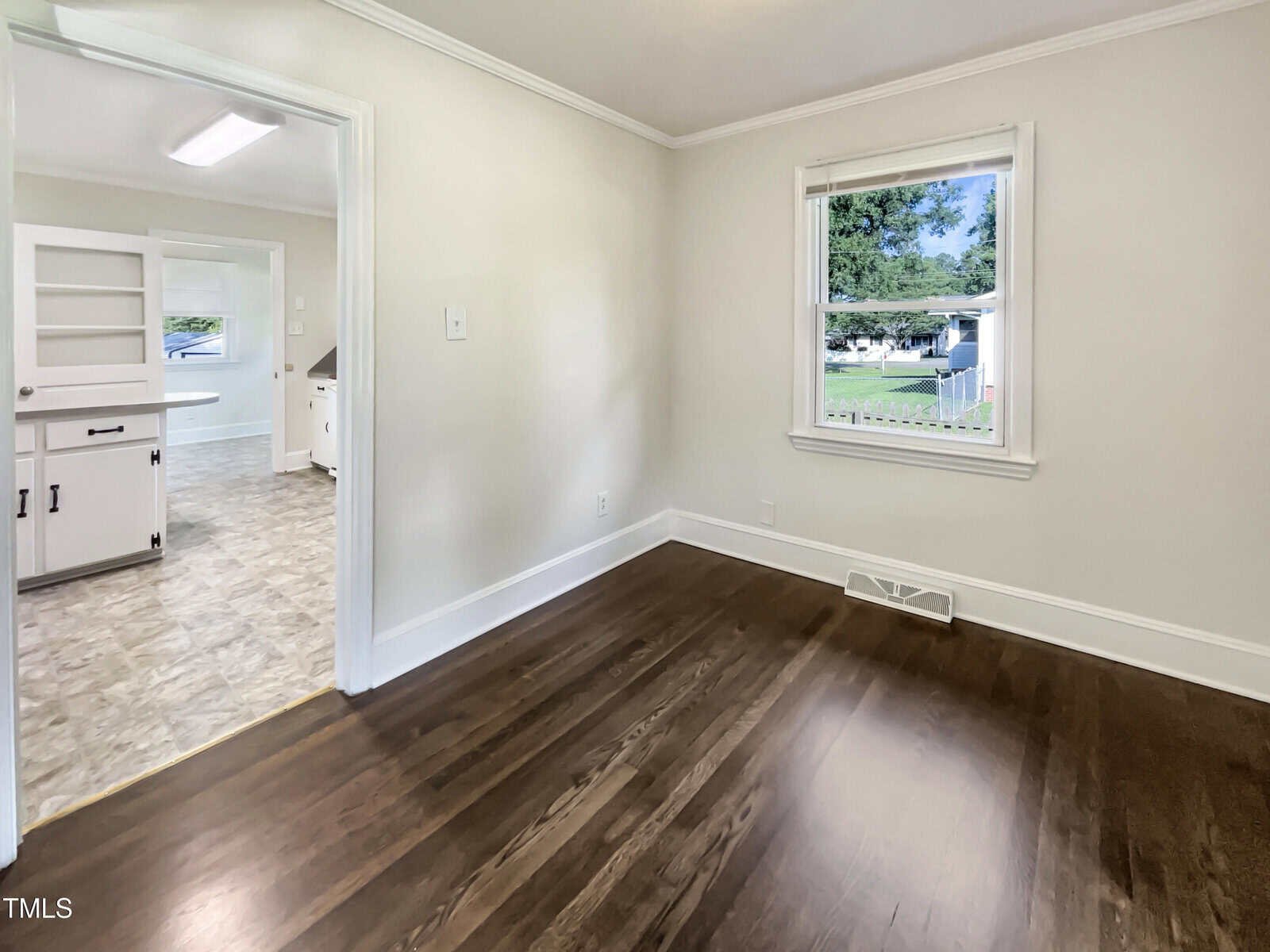 Empty room with dark wood floors, beige walls, white trim, and a vent near the window. An adjoining kitchen with white cabinets and tiled flooring is visible through an open doorway. Daylight shines through the window.