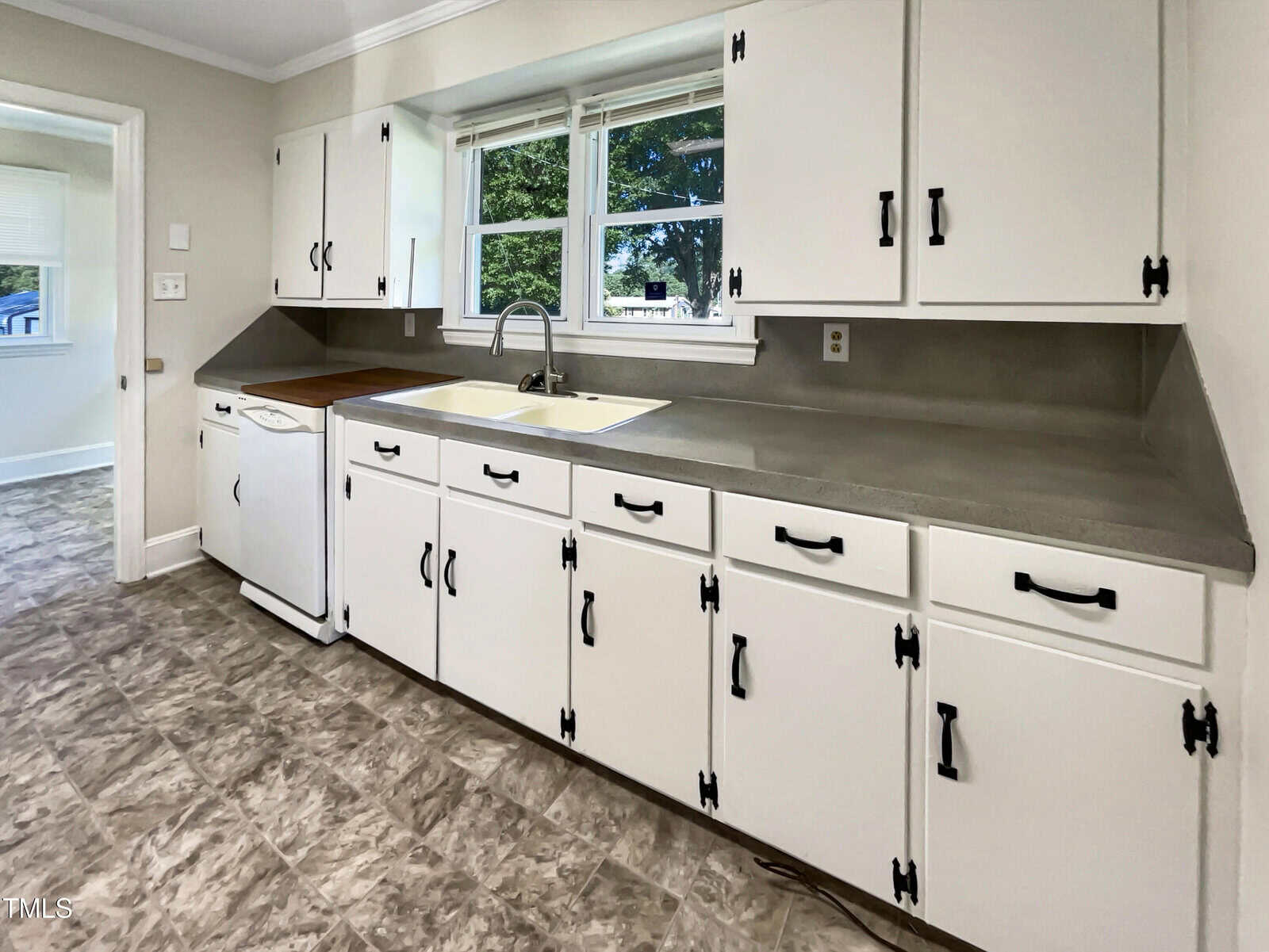 White kitchen cabinets with black handles, a beige countertop, a white sink under double windows, a dishwasher, and gray patterned floor tiles. Bright natural light comes in through the windows.