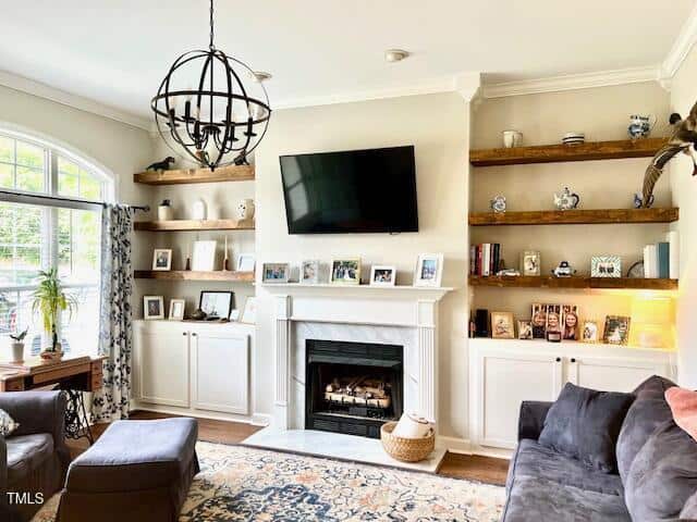 A living room features a wall-mounted TV above a white fireplace, wooden shelves with decor, white cabinets, a large window with curtains, a gray sectional sofa, and a round black chandelier.