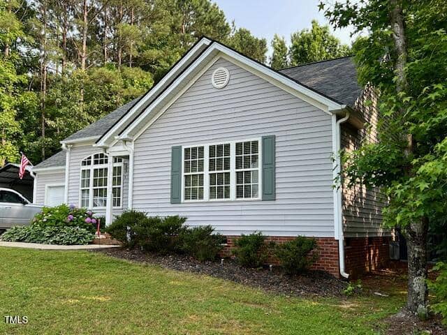 Single-story light gray house with white trim and green shutters, set on a grassy lawn with shrubs and flowers near the front, surrounded by trees.