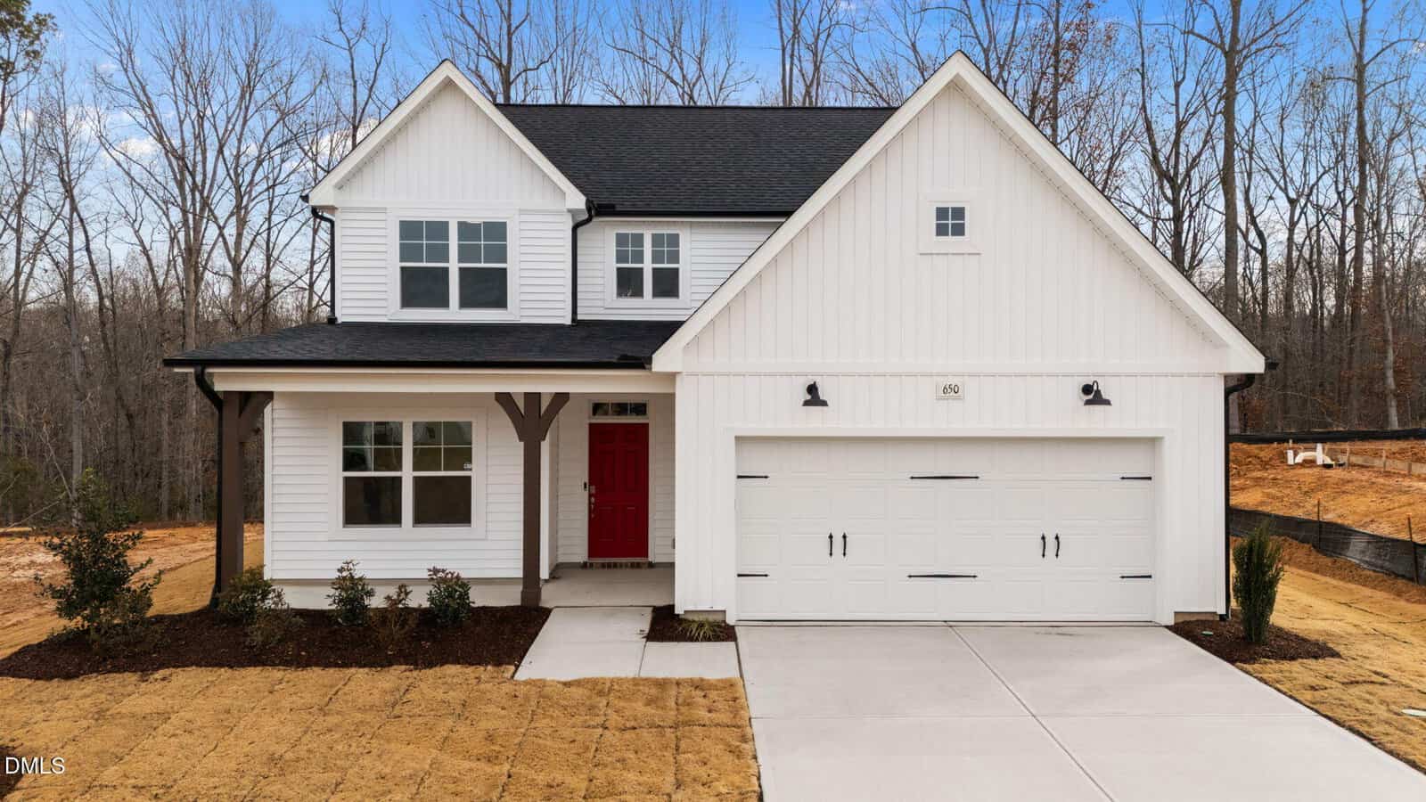 A modern two-story white house with black trim, a red front door, double garage, and newly planted landscaping, situated on a lawn with freshly laid sod and backed by leafless trees.