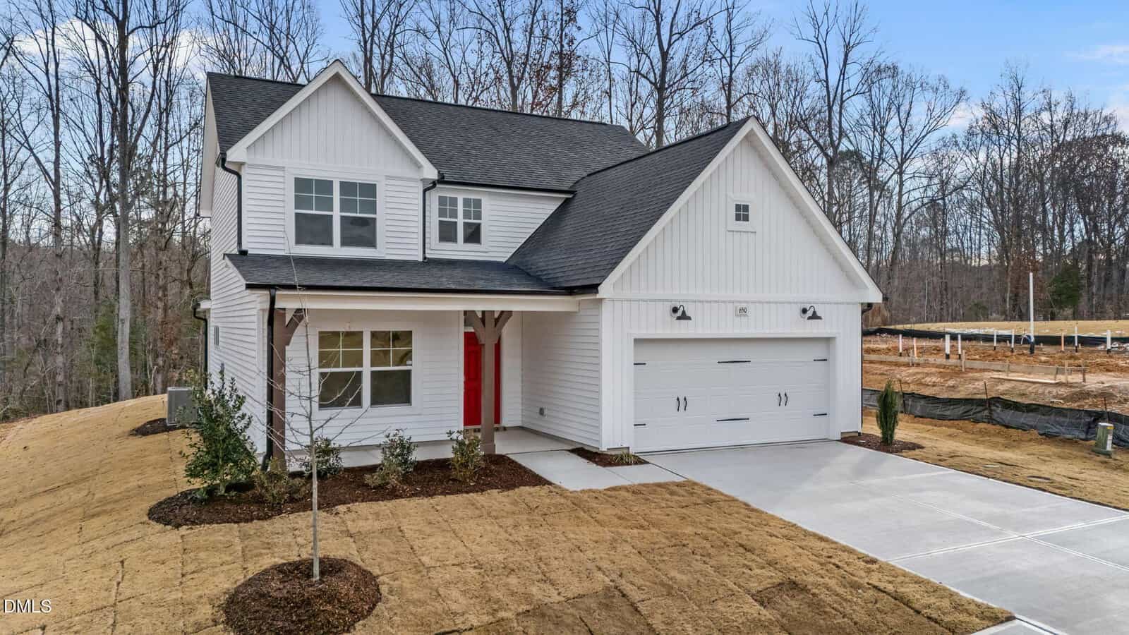 A modern two-story white house with a dark roof, a red front door, and a double garage. The yard has fresh sod and new landscaping, and the house is surrounded by leafless trees and a partially cleared lot.