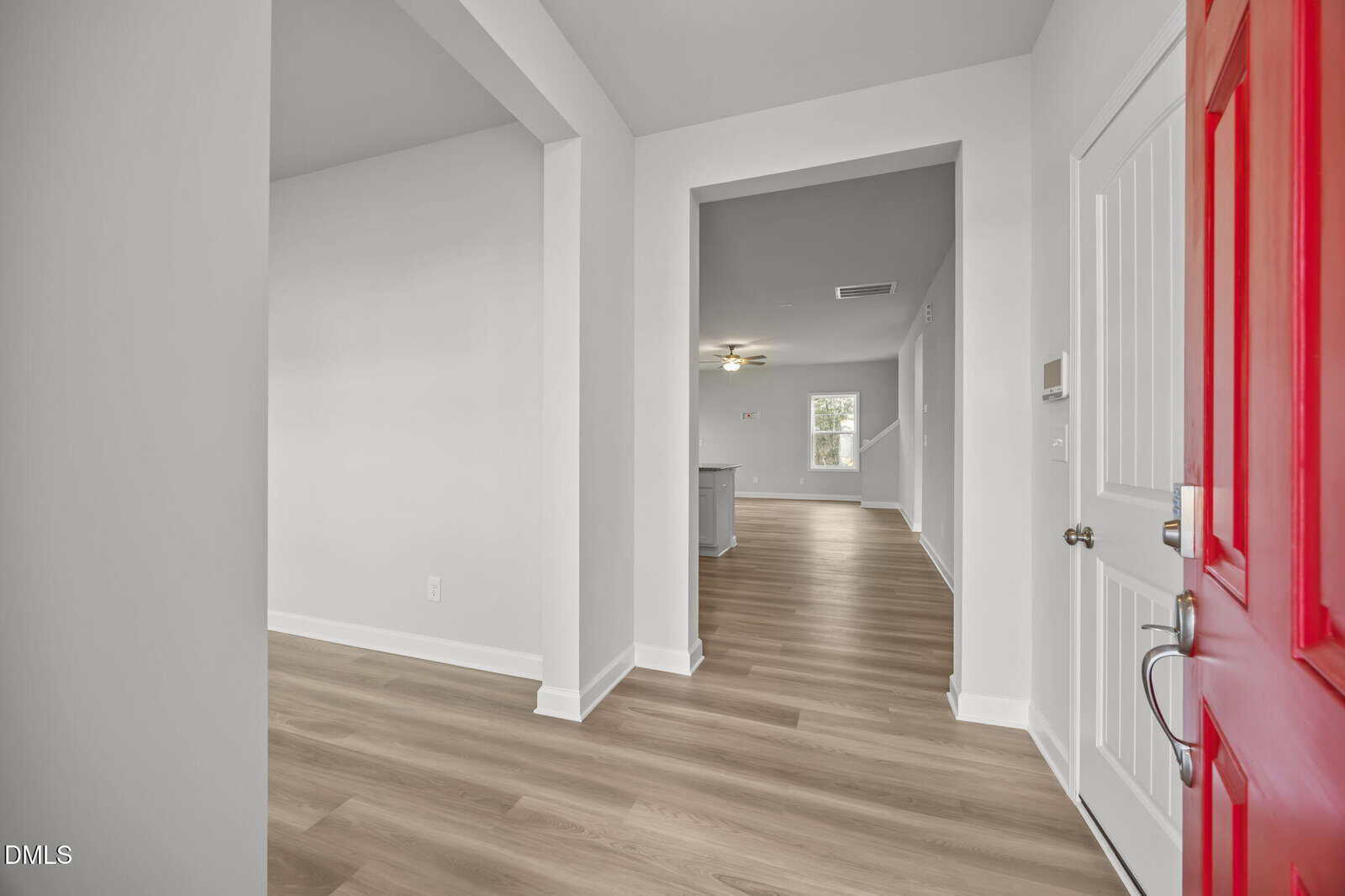View from an open red front door into a modern home with light wood flooring, white walls, and an open hallway leading to a bright living area and kitchen in the background.