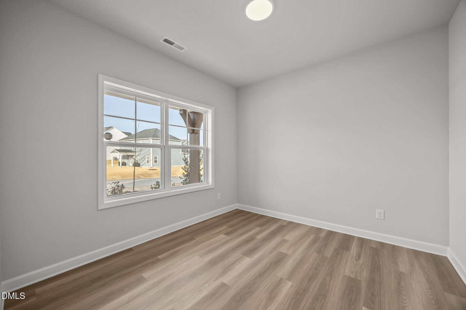 Empty room with light wood flooring, light gray walls, a large window letting in natural light, and a view of neighboring houses and yard outside. Ceiling light and baseboard visible.