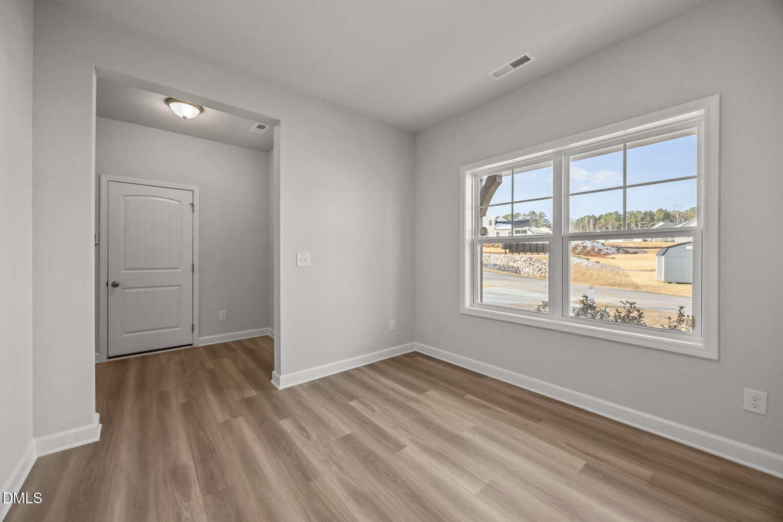 A bright, empty room with light wood flooring, white walls, a large window letting in natural light, and a closed white door in a small hallway area.