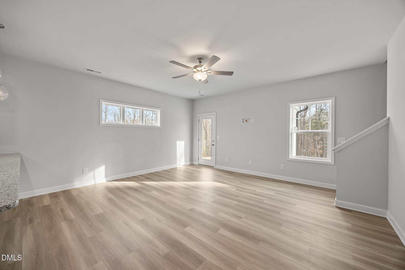 Bright, empty living room with light wood floors, white walls, ceiling fan with light, large windows, and a door leading outside. Stairs are visible to the right.