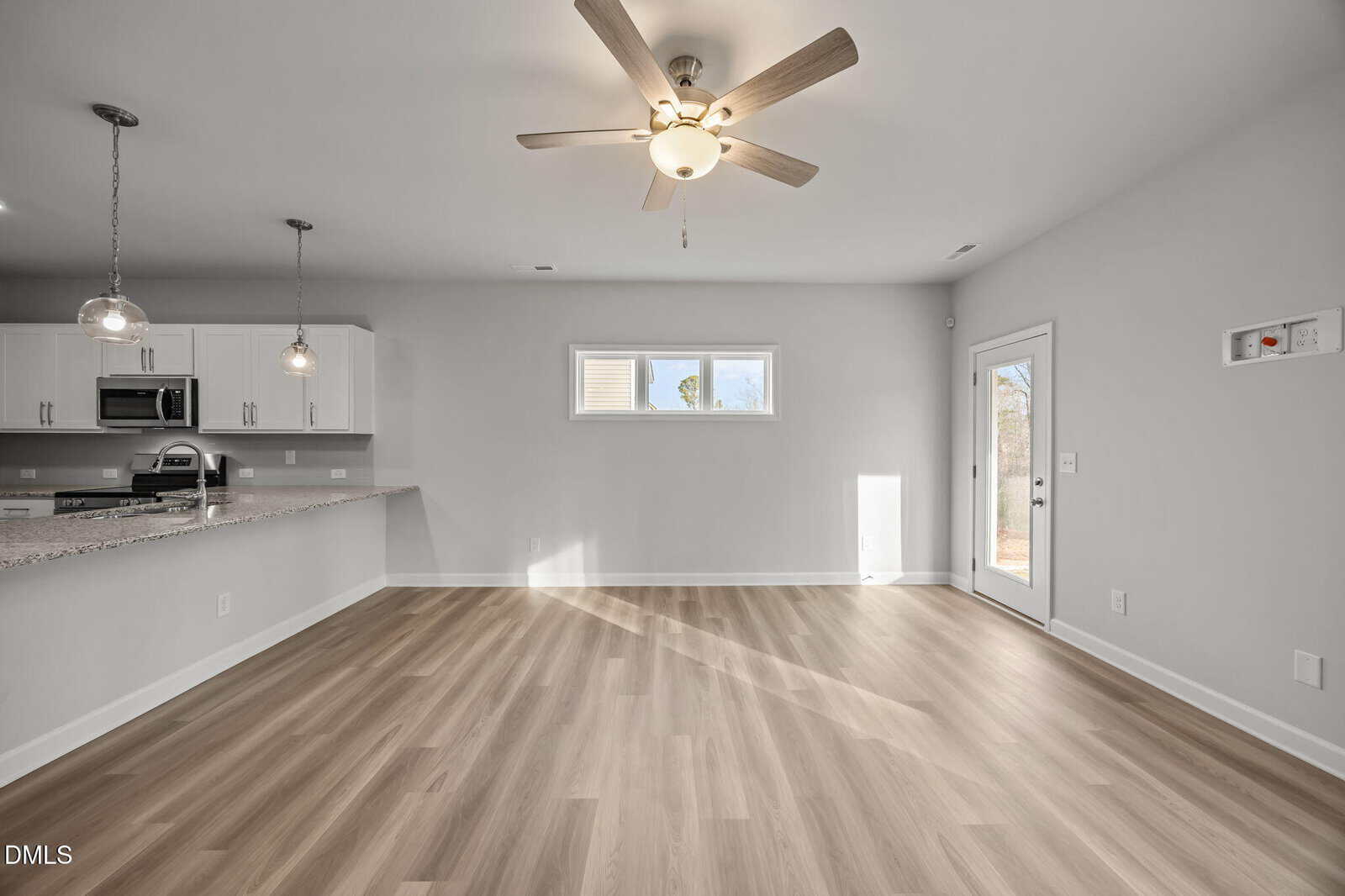 Bright, empty room with light wood floors, ceiling fan, and recessed lighting. Open kitchen with white cabinets and granite counters. Two pendant lights above the counter. Small window and a glass door let in natural light.