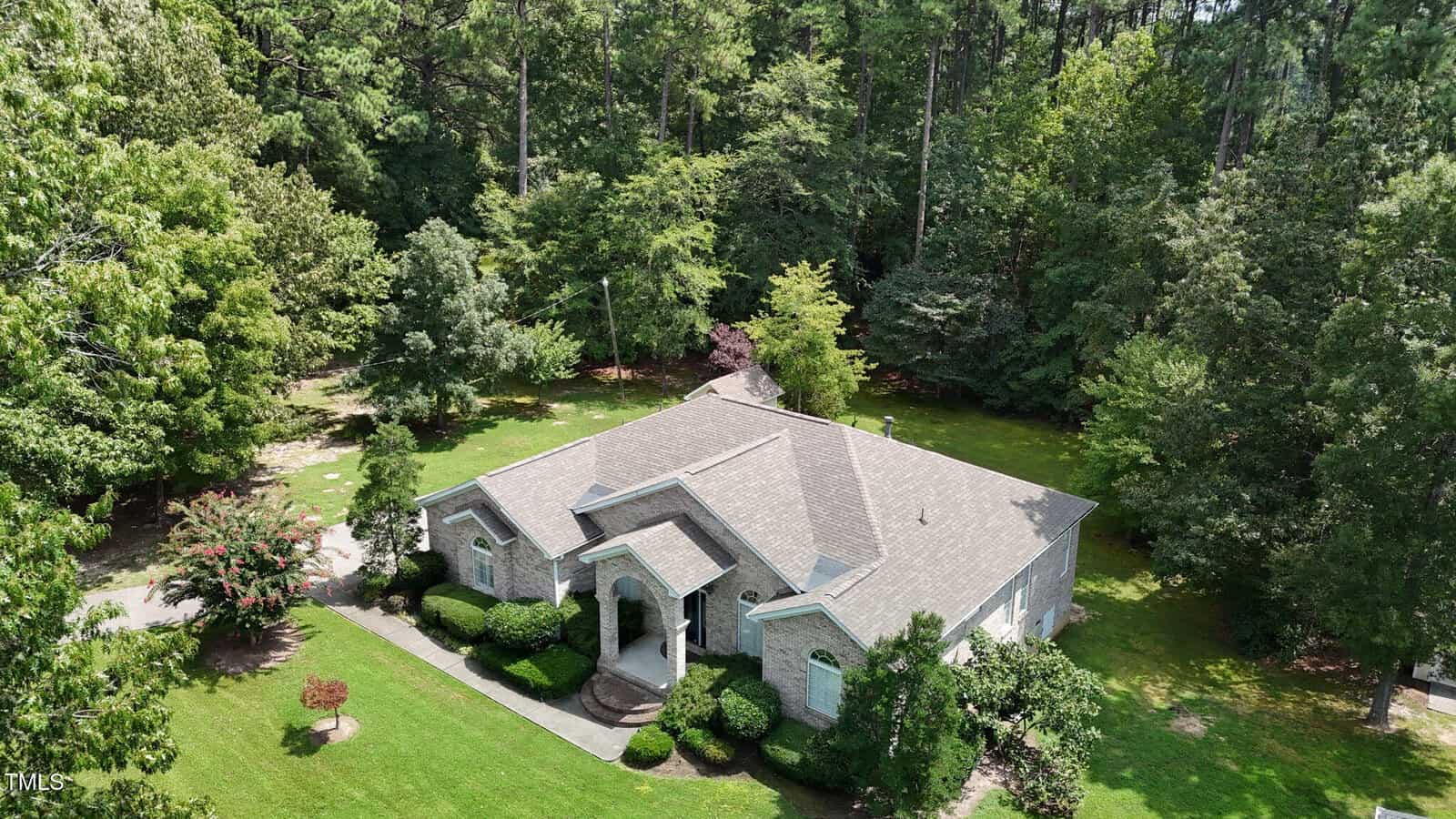 Aerial view of a single-story gray brick house with a gabled roof, surrounded by green lawn, shrubs, and dense trees under a clear sky. A covered entryway is visible at the front of the house.
