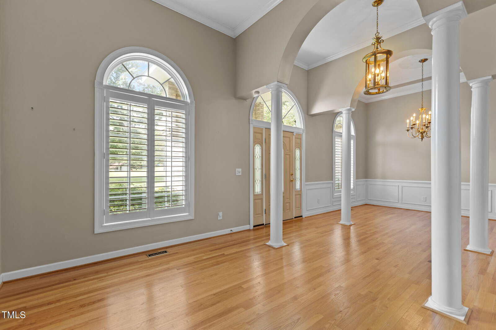 Spacious entryway and dining area with hardwood floors, large arched windows with white shutters, double front doors, high ceilings, white columns, and gold chandeliers.