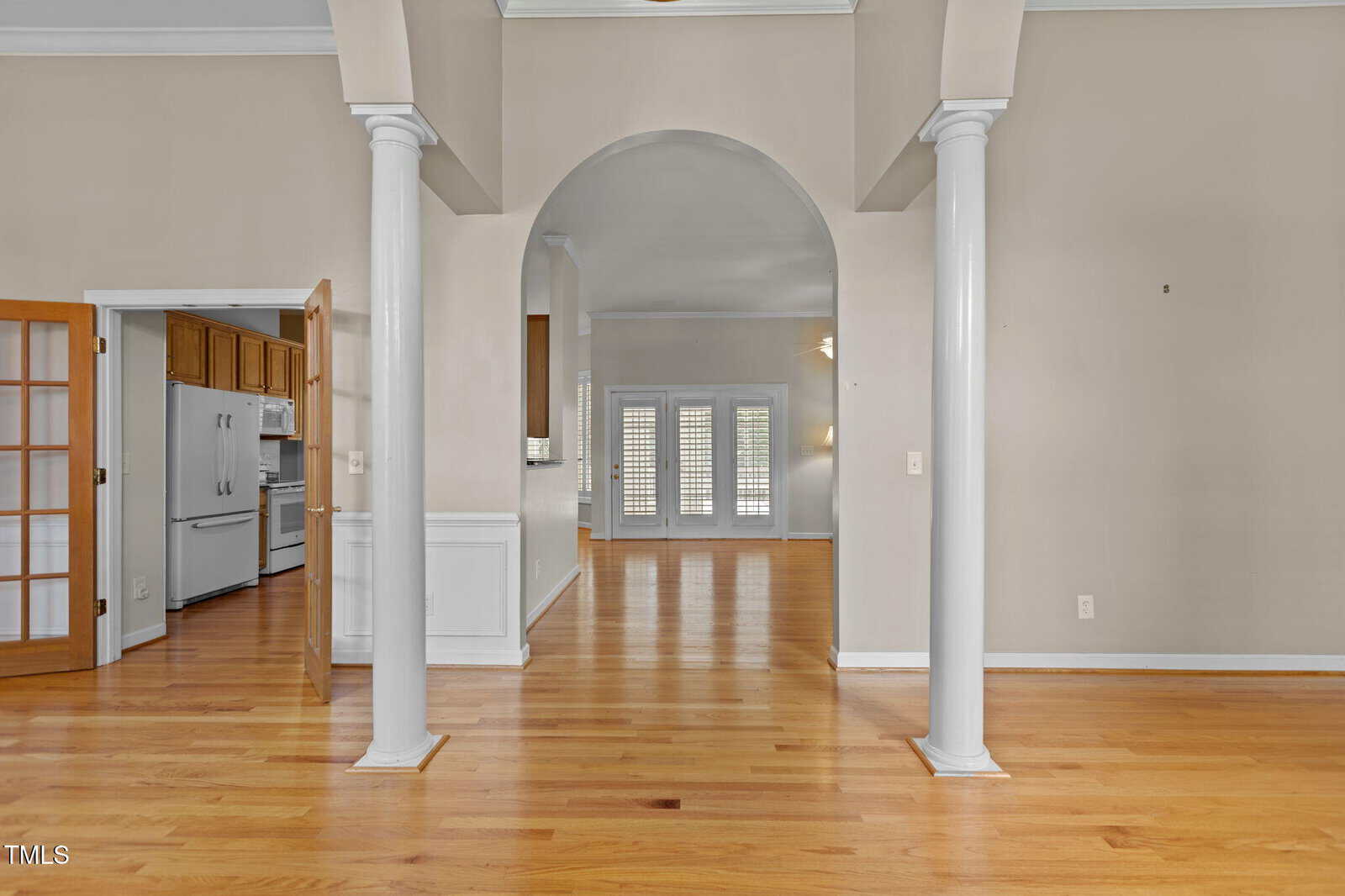 Open indoor space with light wood floors, beige walls, and white columns. An arched doorway leads to another room with double doors and shutters. A kitchen with wooden cabinets is visible to the left.
