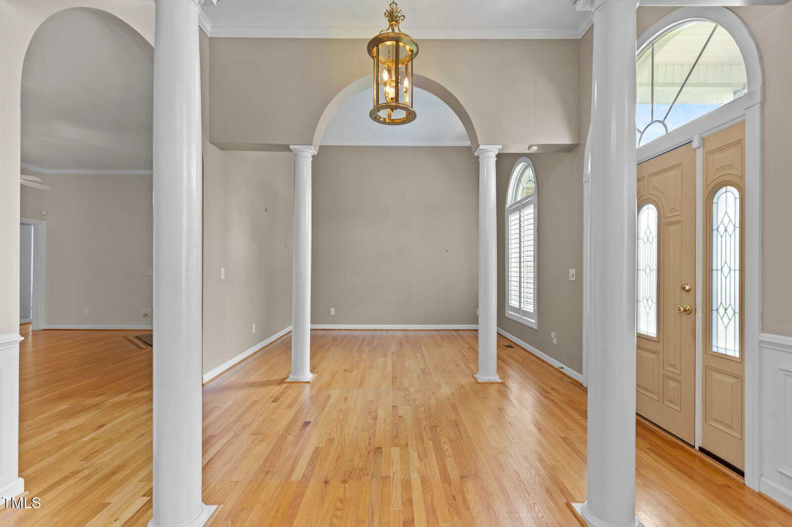 Interior view of a foyer with light wood flooring, beige walls, white columns and trim, an arched entryway, and a double front door with glass panels. A hanging lantern light fixture is centered above.