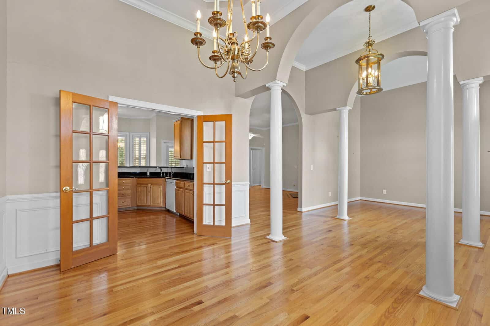 A spacious open room with hardwood floors, tall white columns, beige walls, French doors leading to a kitchen with wooden cabinets, and gold chandeliers hanging from the ceiling.