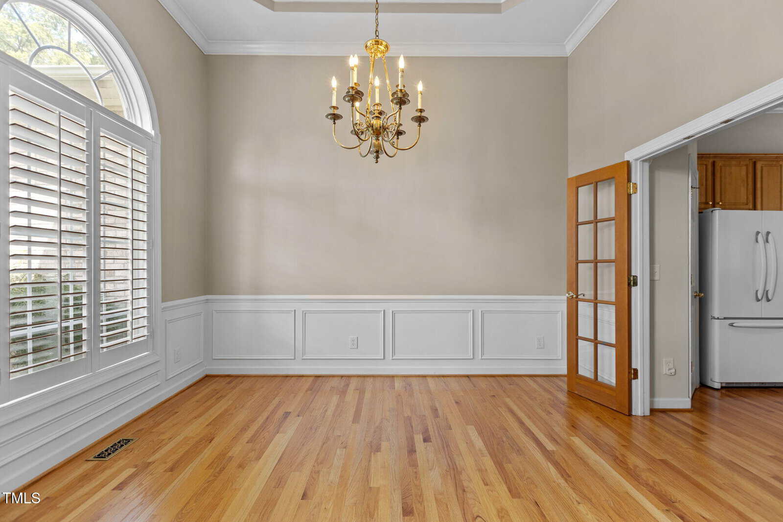 A dining room with hardwood floors, beige walls, white wainscoting, a gold chandelier, large arched window with shutters, and an open French door leading to a kitchen with a white refrigerator.