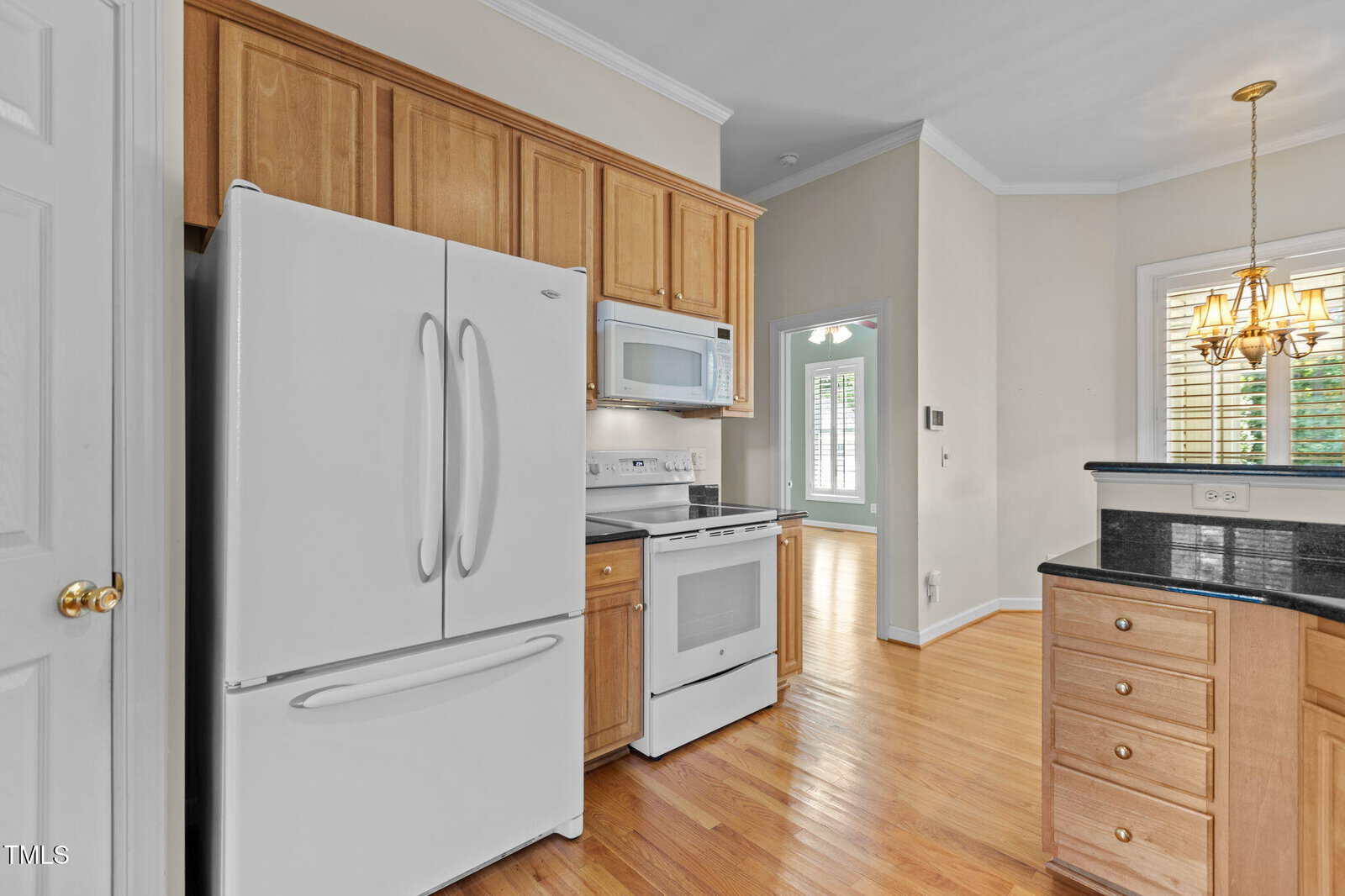 A kitchen with wood cabinets, white appliances including a refrigerator, stove, and microwave, granite countertops, and hardwood flooring. A doorway leads to a hallway with a window and a hanging light fixture.