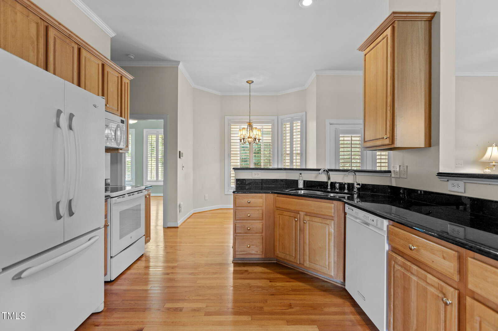 A kitchen with light wood cabinets, black granite countertops, white appliances, a double sink, hardwood floors, and a dining area with large windows and a chandelier in the background.